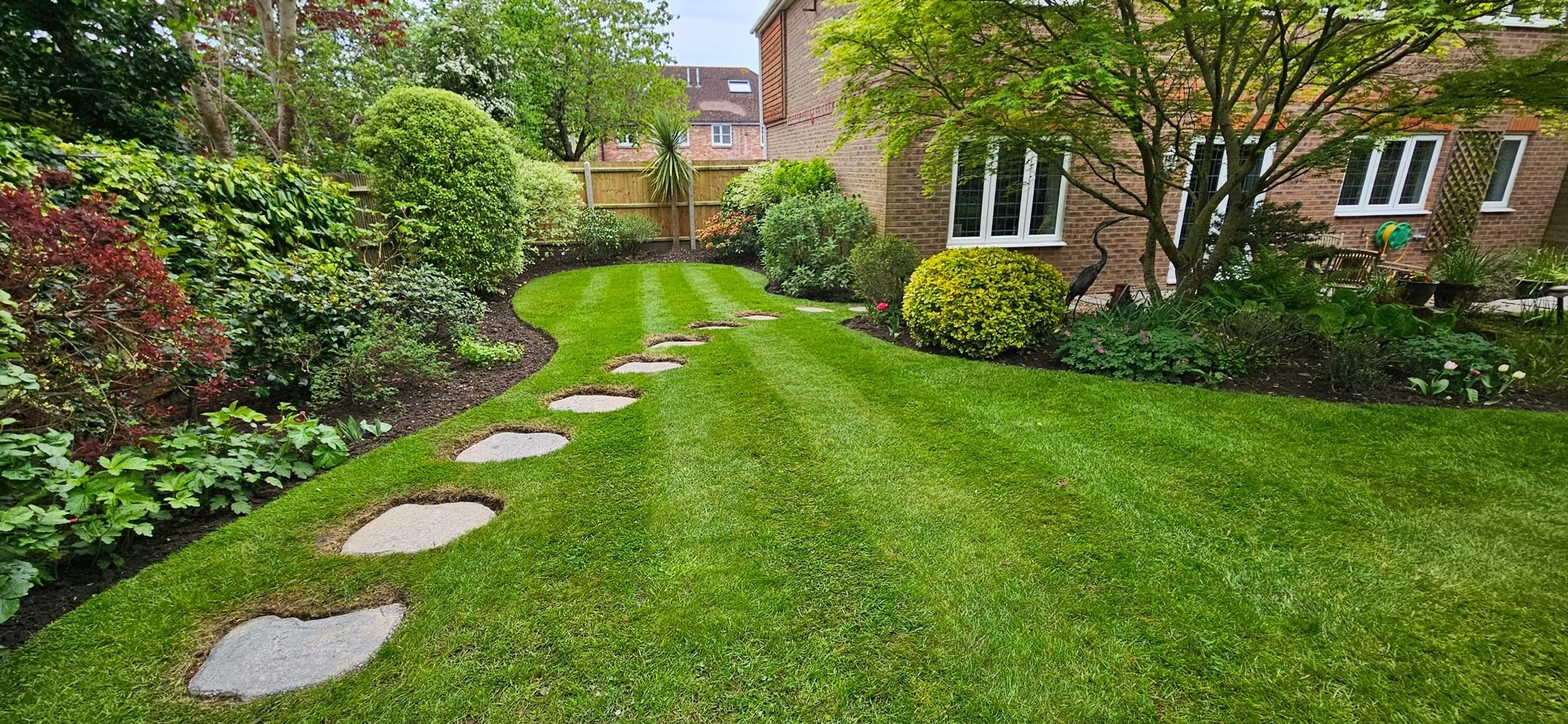 A well-maintained backyard garden with a curved lawn, stepping stones, various shrubs, flowers, and trees, with a brick house in the background and a wooden fence.