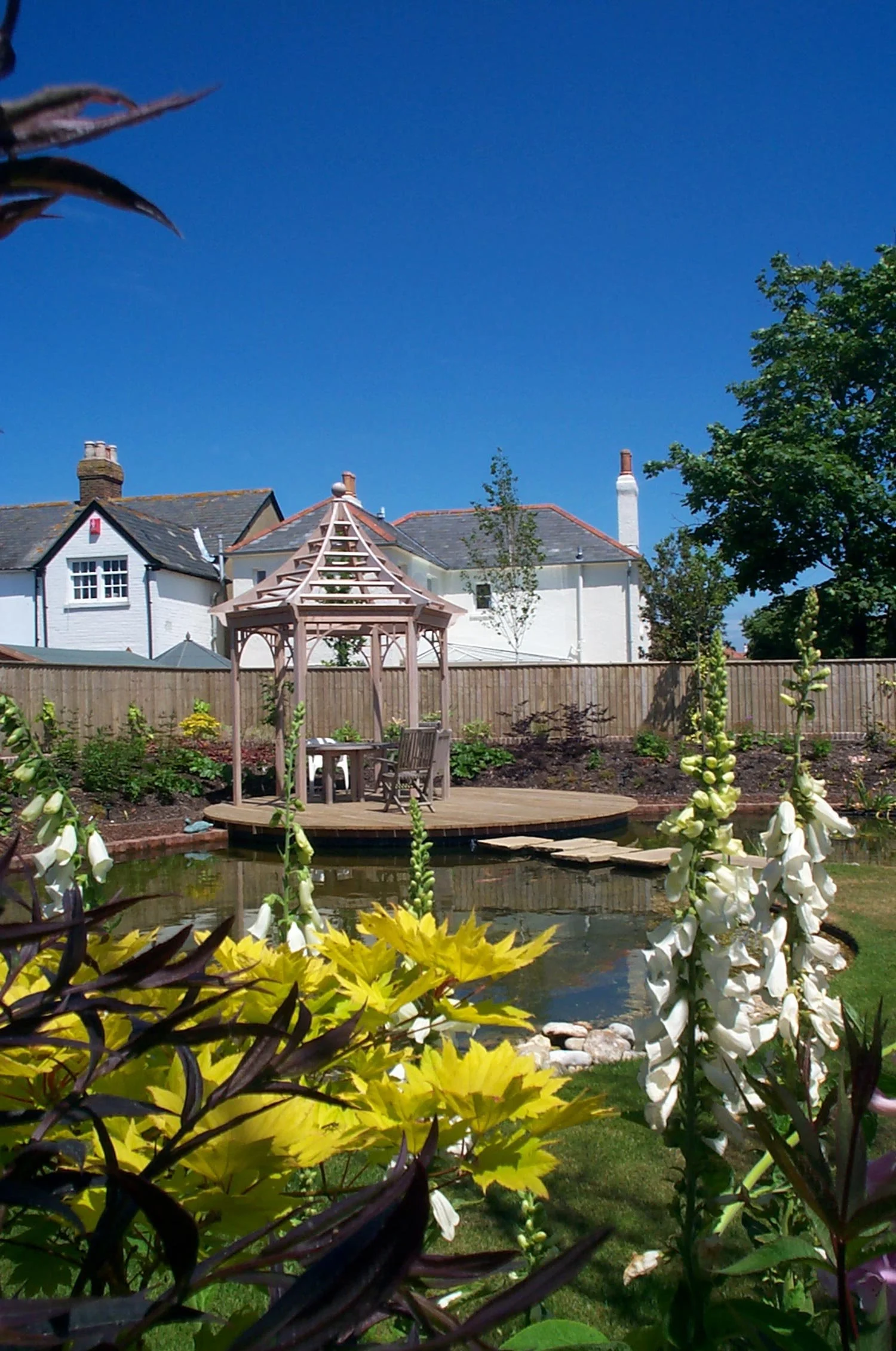 A peaceful backyard garden with a small pond, a wooden gazebo, and blooming flowers. Houses and a blue sky are visible in the background.