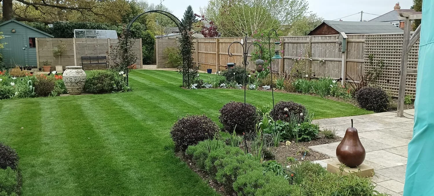 A well-maintained backyard garden with a lush green lawn, flower beds, shrubs, a small patio area with a pear-shaped sculpture, and a garden shed in the background surrounded by a wooden fence.