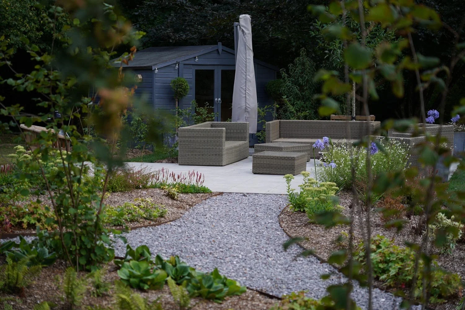 A backyard patio area with wicker furniture, including a sectional sofa and chairs, surrounded by plants and flowers, with a small shed or garden house in the background.