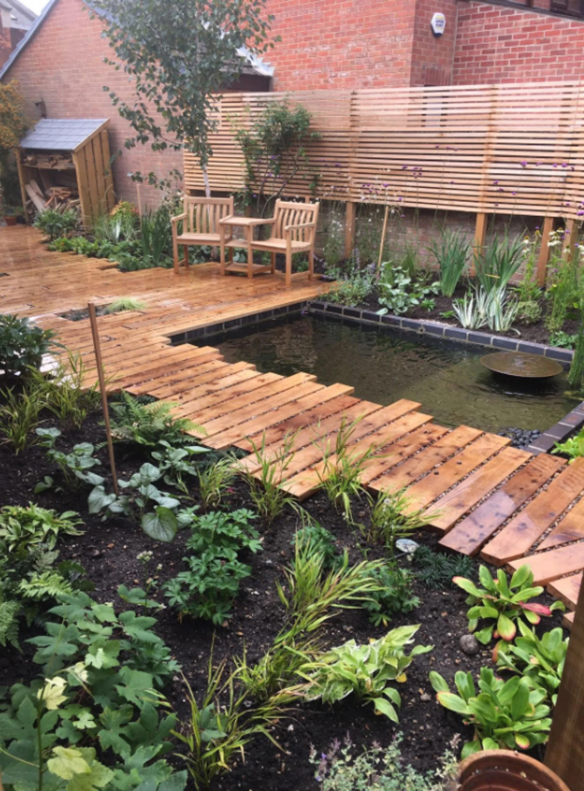 A backyard garden with a small pond, wooden stepping stones, a new wooden deck with two chairs and a small table, surrounding lush green plants, and a wooden fence in the background.
