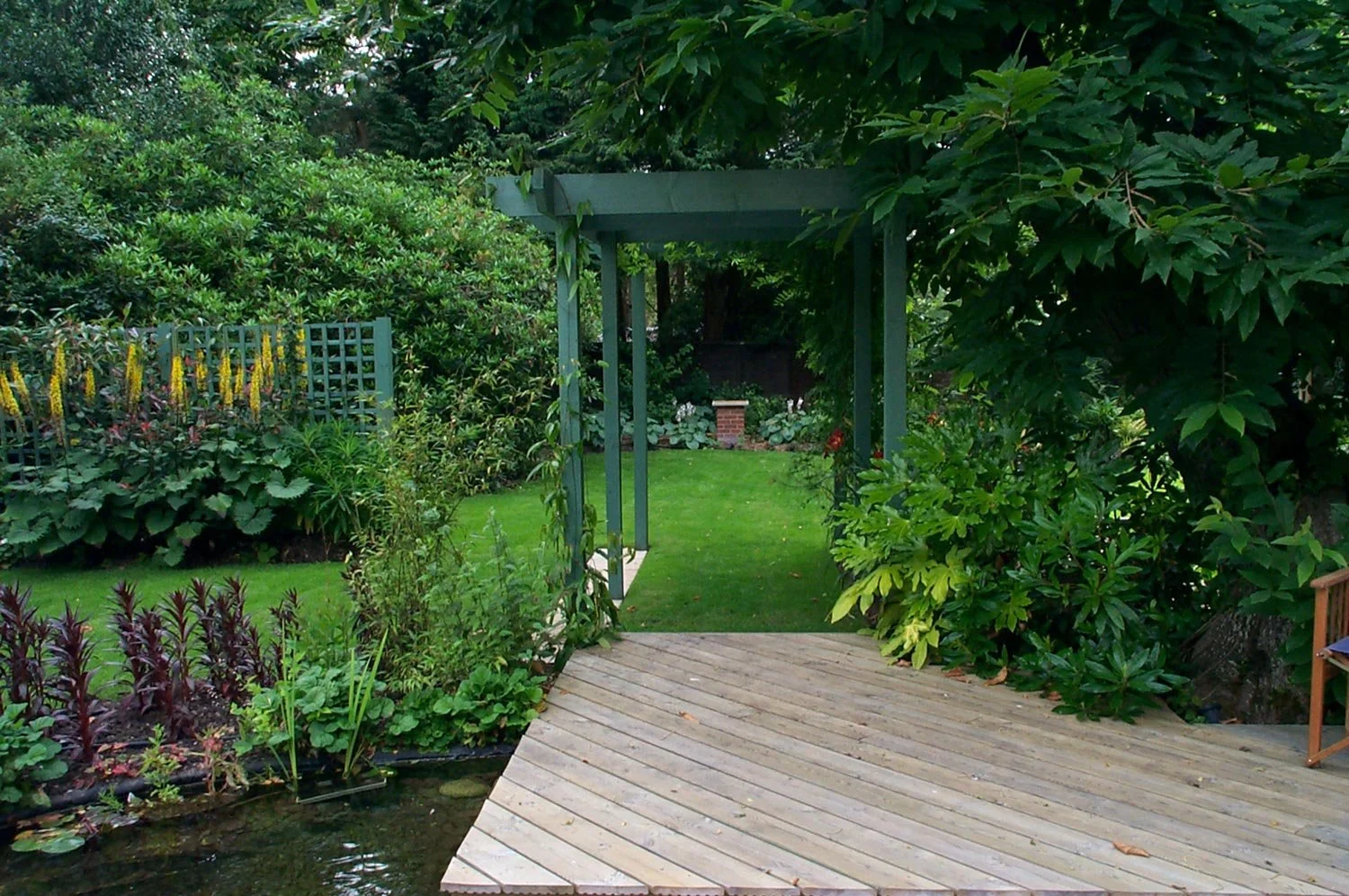A lush green garden with a wooden deck, a small pond, and a green arbor over a grassy pathway. Dense foliage and flowering plants surround the area.