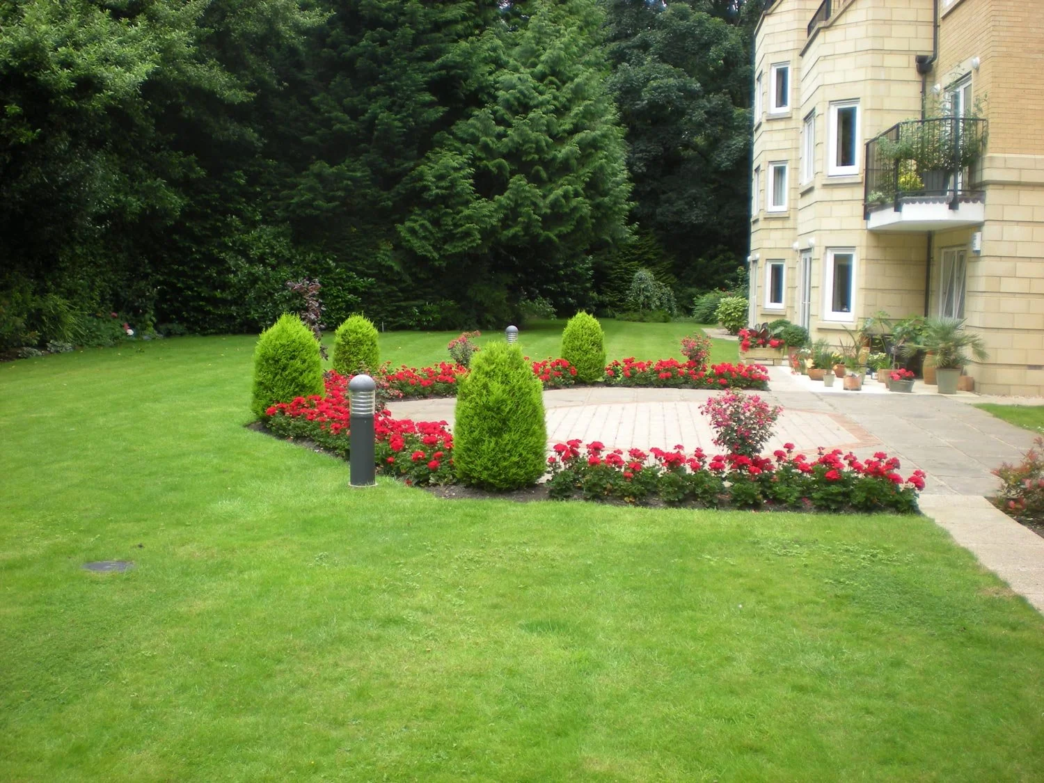 A well-maintained garden with a green lawn, small bushes, and a flower bed with red flowers surrounding a paved area, adjacent to a beige apartment building with potted plants.
