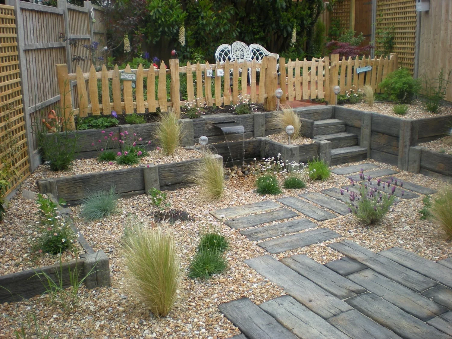A small, landscaped backyard garden with gravel ground cover, wooden edging, stone steps, a small waterfall, and plants including ornamental grasses and blooming flowers. A white decorative bench is in the background, enclosed by a wooden picket fenc