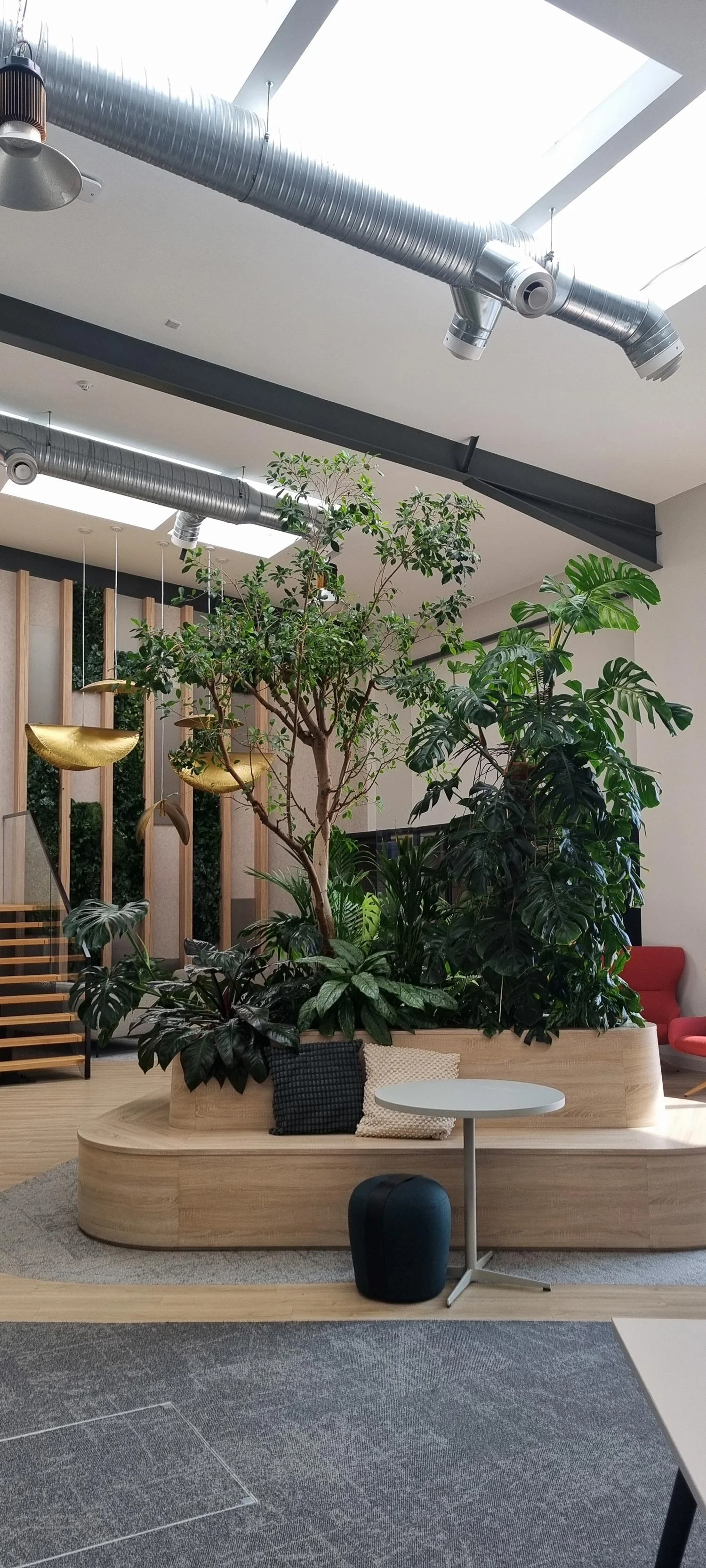 Indoor space with large planter containing trees and lush green plants, a white round table, a black ottoman, and a red chair in the background. Modern decor with suspended gold decorative pieces and industrial ceiling ducts.