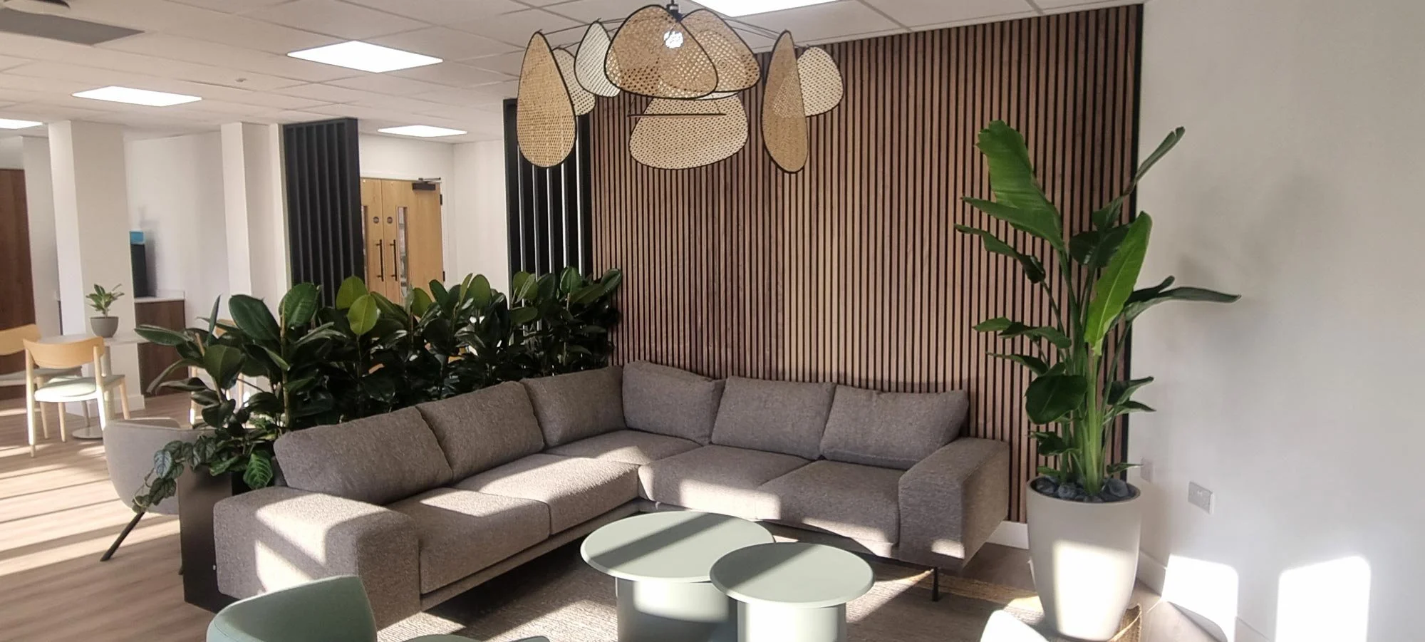 Modern hotel lobby with beige L-shaped sofa, green plants, round coffee tables, wooden slat wall, hanging woven light fixtures, and light-colored walls and flooring.