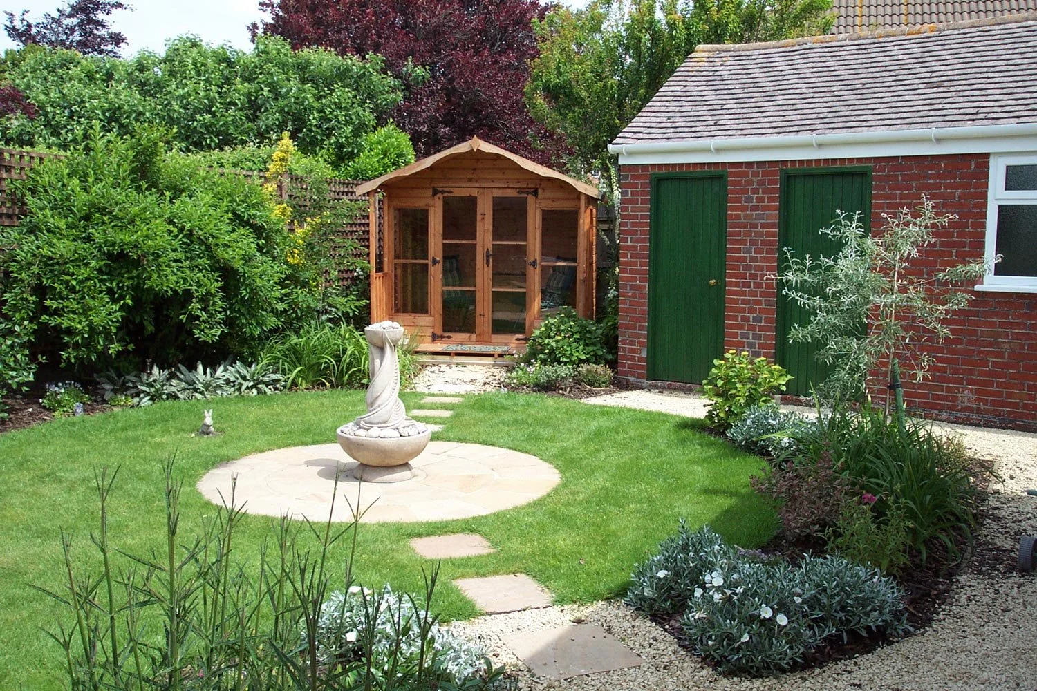 A well-maintained backyard garden with green grass, a small rabbit statue, and a stone birdbath as central features. There is a wooden garden shed with glass doors in the background, surrounded by lush shrubs and trees. To the right, a brick building