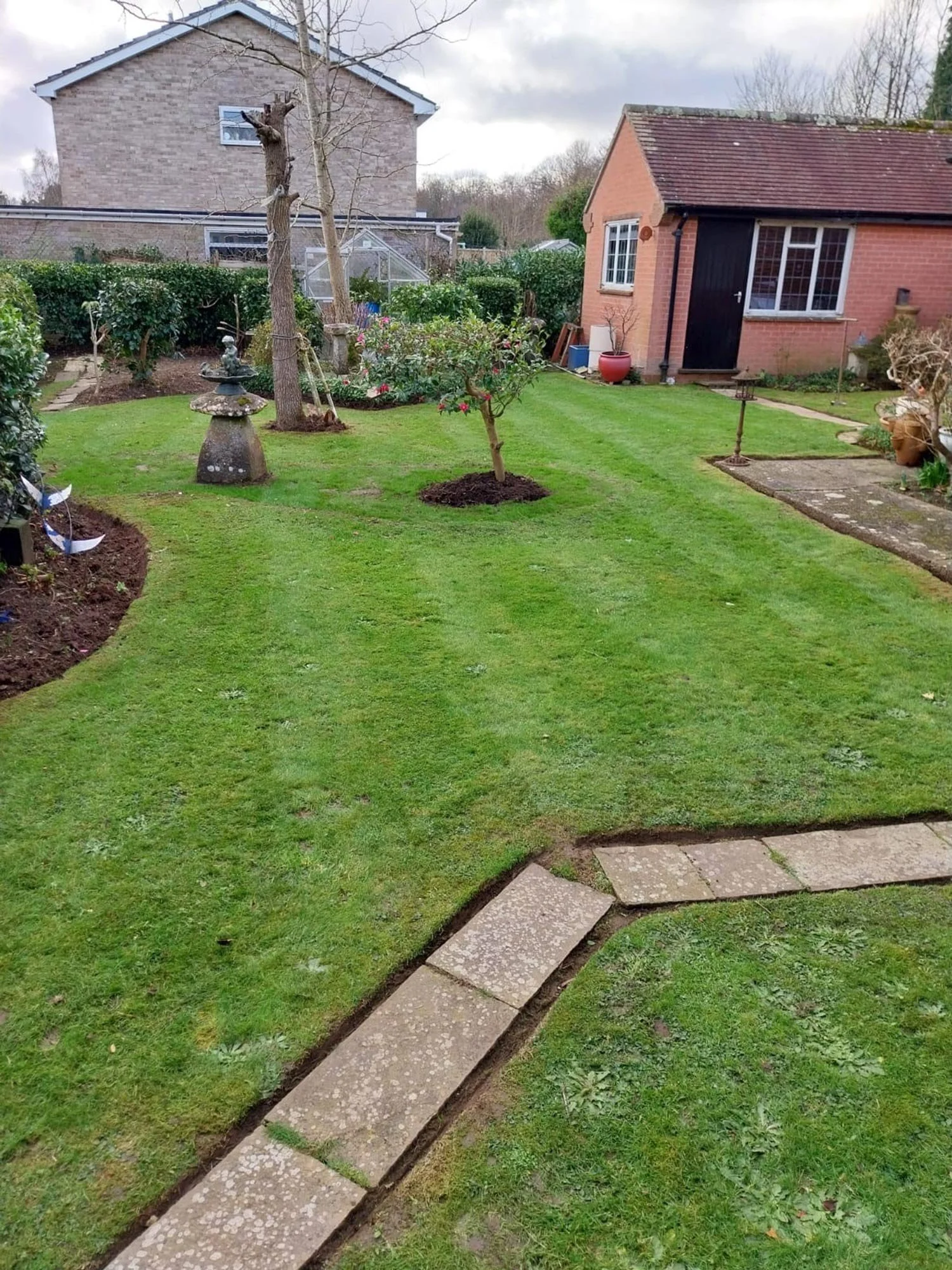 A backyard with a well-maintained lawn, small trees, garden beds, and garden ornaments, including a stone lantern and birdbaths. There is a brick structure with a black door and window, and a larger house in the background. The sky is cloudy.