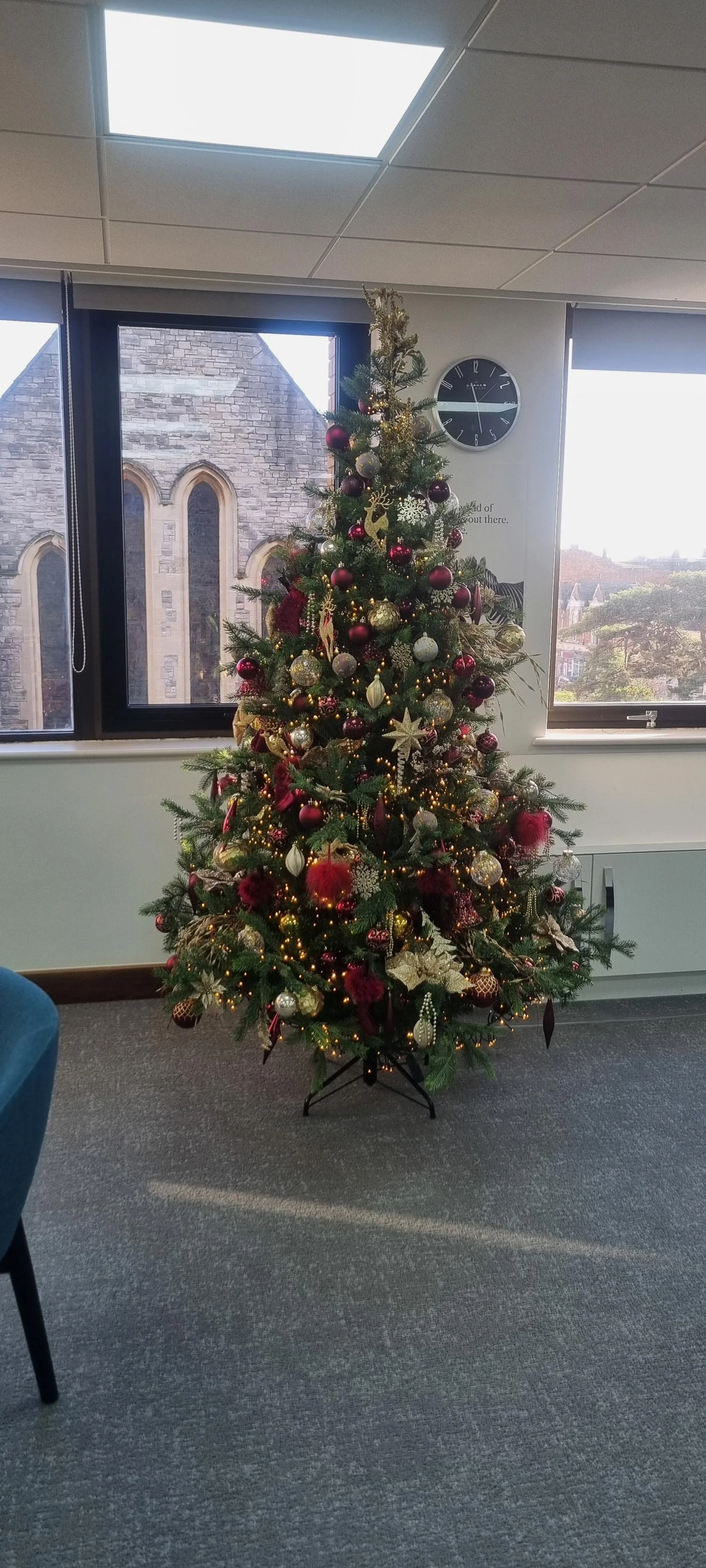 Decorated Christmas tree with red, gold, and silver ornaments, golden stars, and string lights indoors near windows.