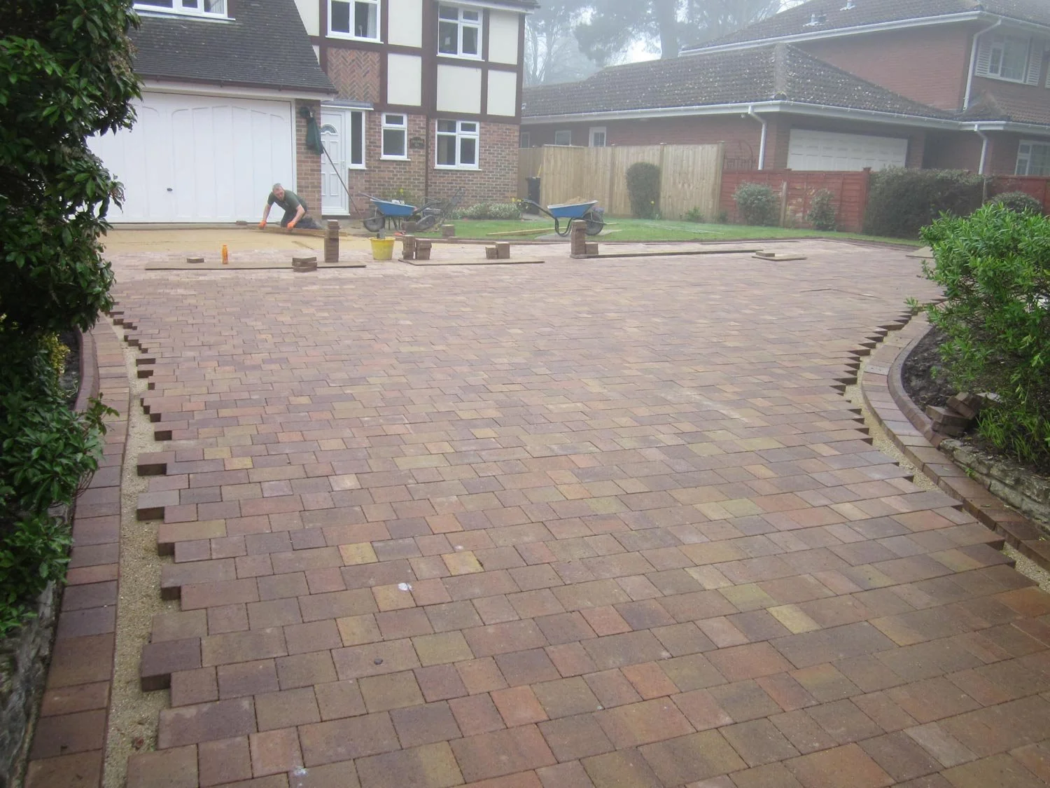 A person working on installing or repairing a brick driveway in a residential area with houses, trees, and bushes in the background.