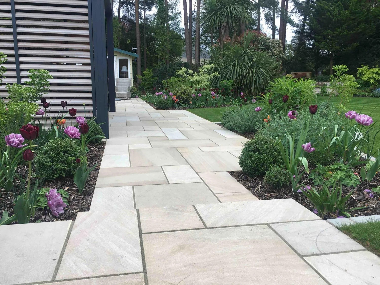 A garden pathway with light-colored stone tiles, bordered by blooming purple and dark red flowers, leading past a dark panel wall toward a garden shed surrounded by trees and lush greenery.