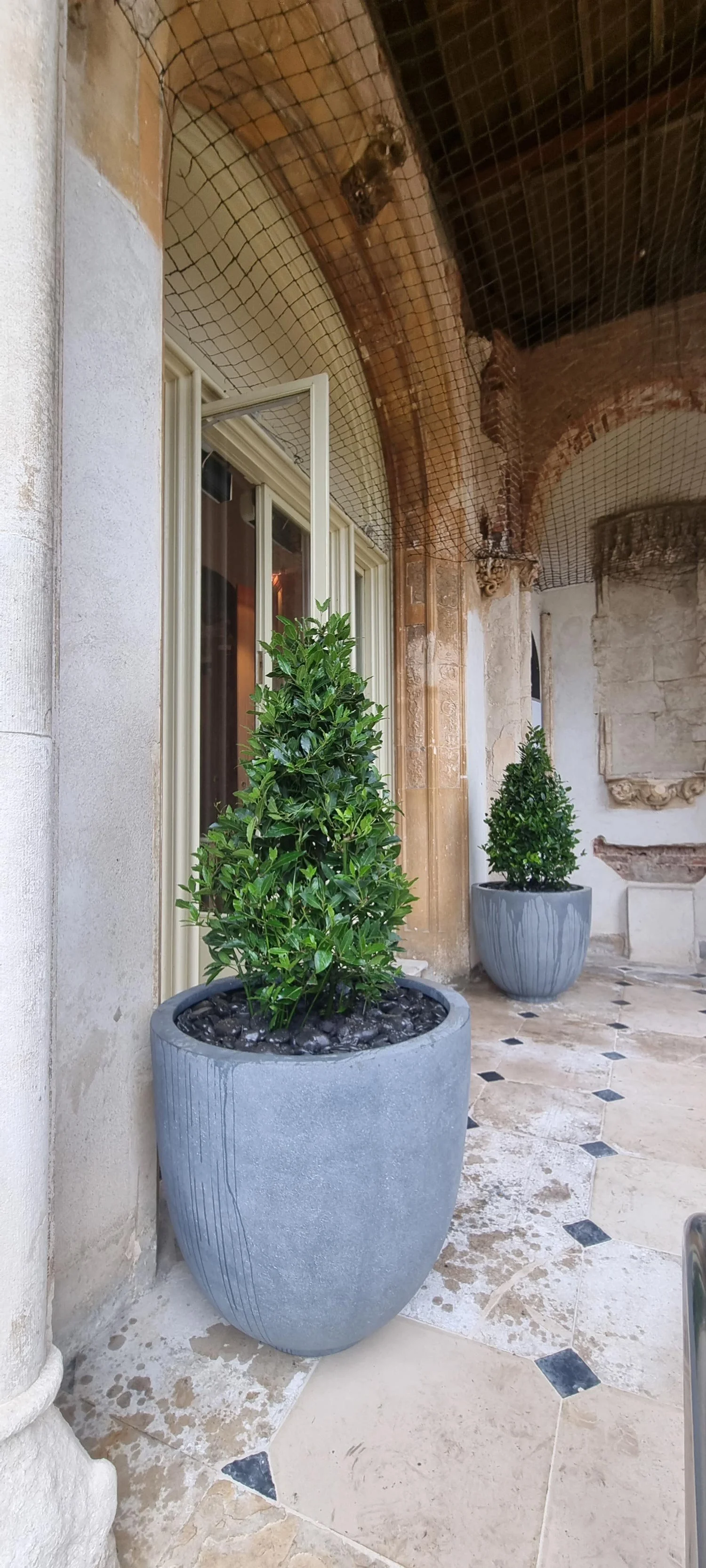 Two large gray planters with green shrubs on an outdoor patio with stone flooring and ornate architecture.