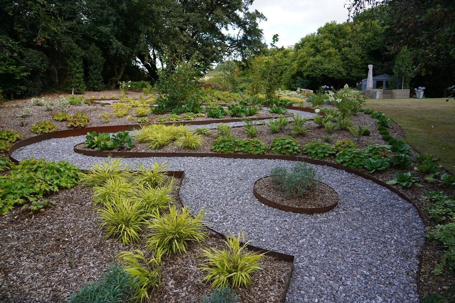 A landscaped garden with a winding gravel pathway, various green plants, and small shrubs, surrounded by trees and a grassy area with outdoor furniture and decorative planters in the background.