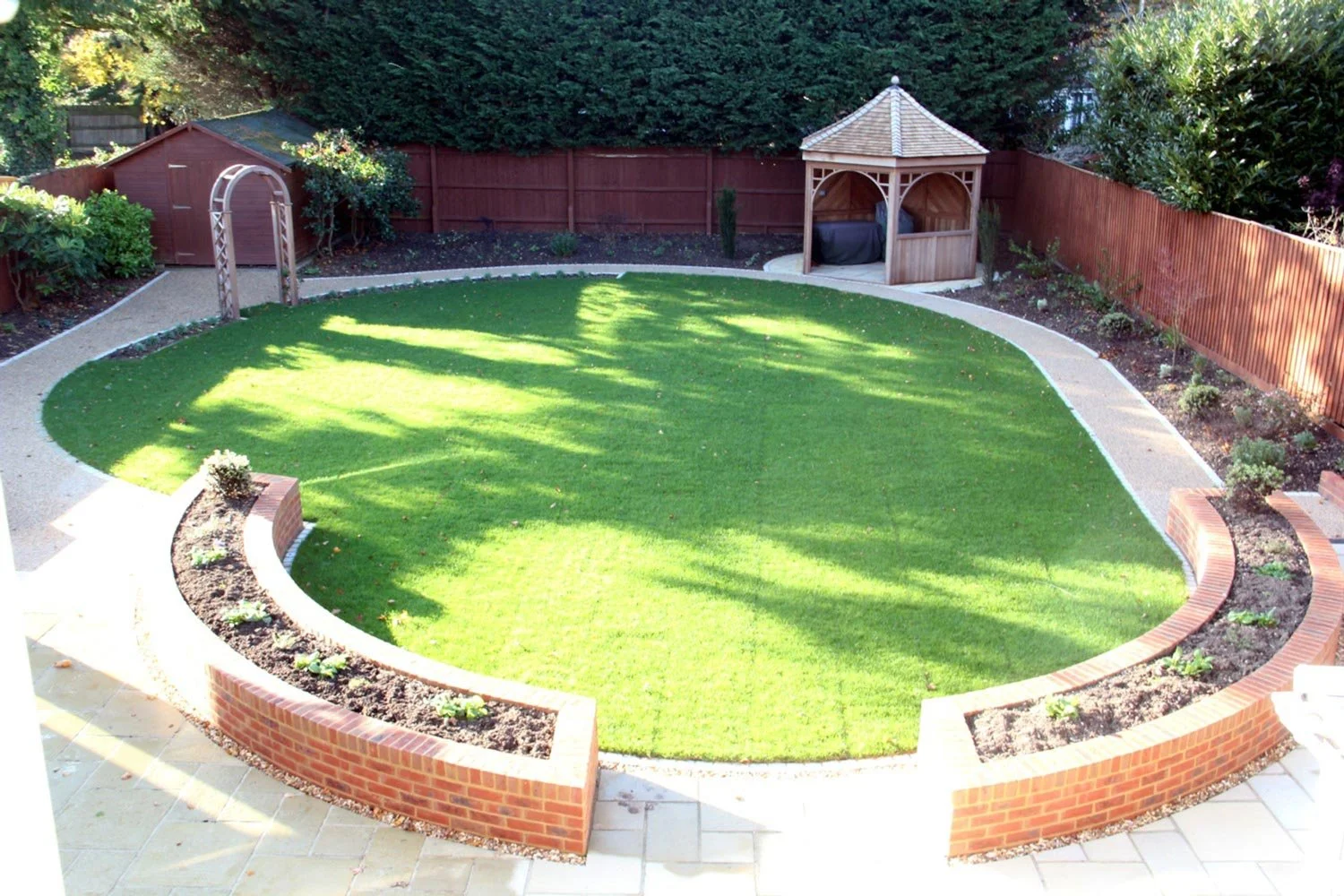 A backyard with a well-maintained green lawn, edged by curved brick and stone borders, including a flower bed, a wooden garden shed, a wooden gazebo, and a surrounding wooden fence.