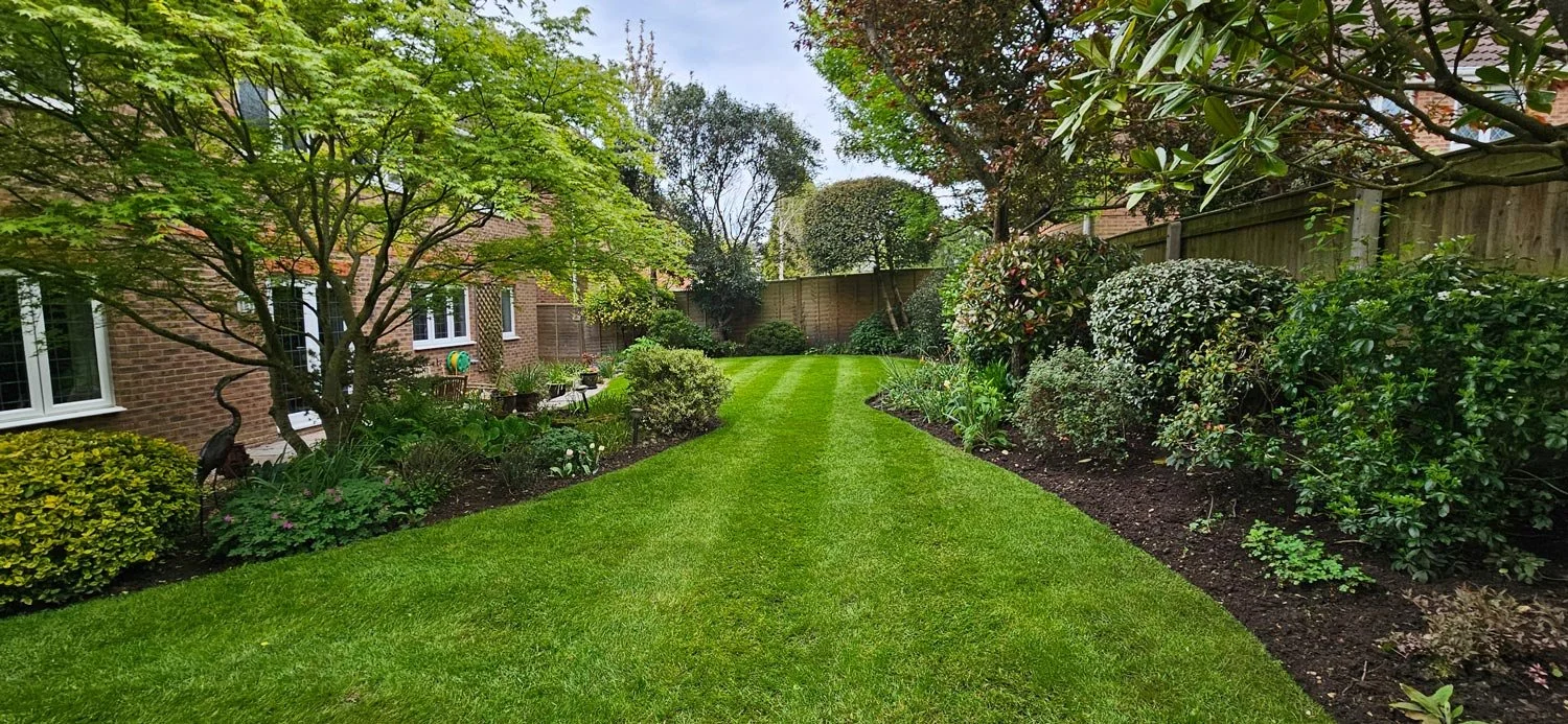 A well-maintained backyard garden with lush green grass, trees, shrubs, and flower beds, adjacent to a brick house with white-framed windows and a wooden fence.