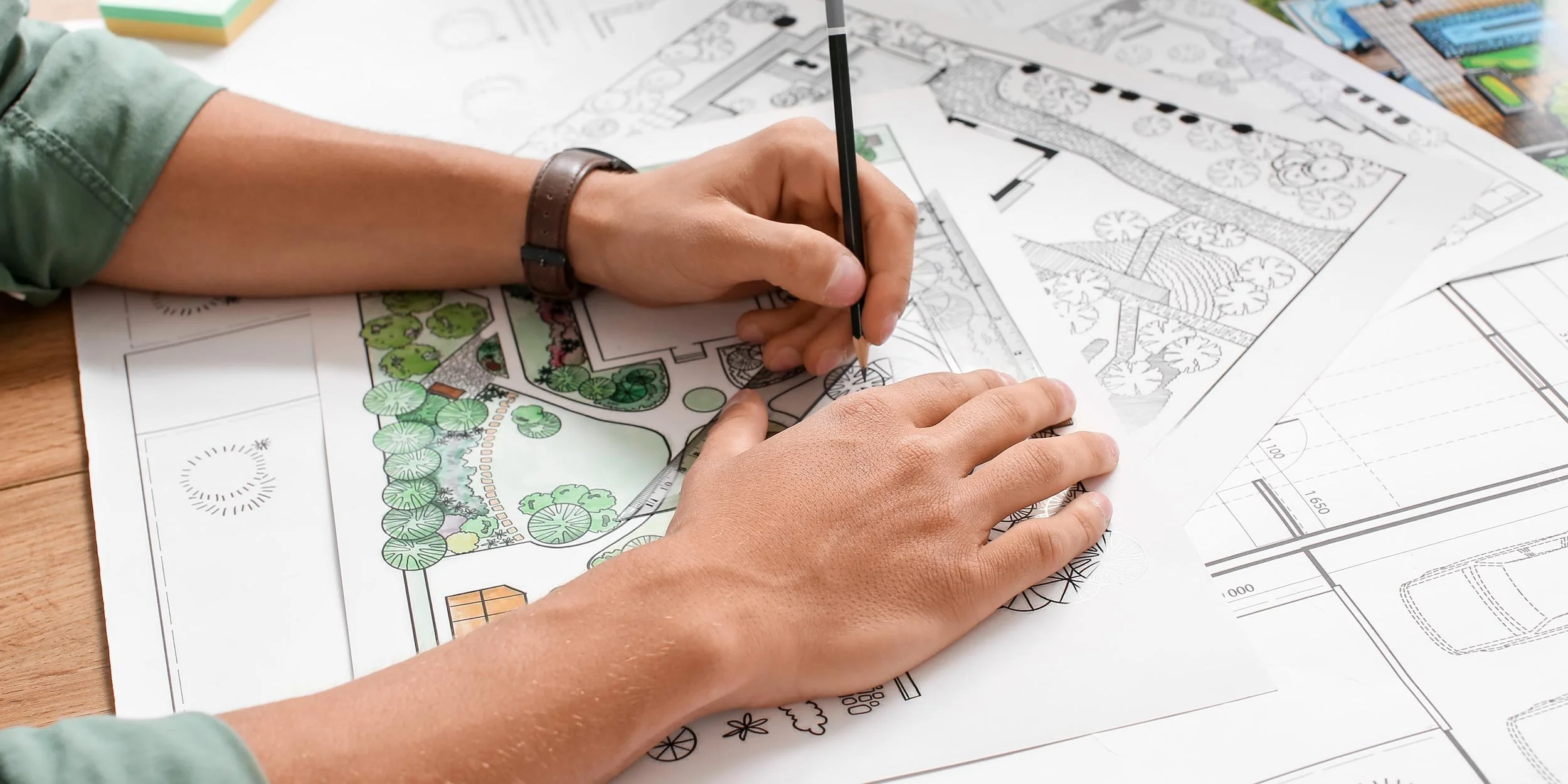 Person sketching and coloring landscape plans on architectural drawings with trees, pathways, and layout details.