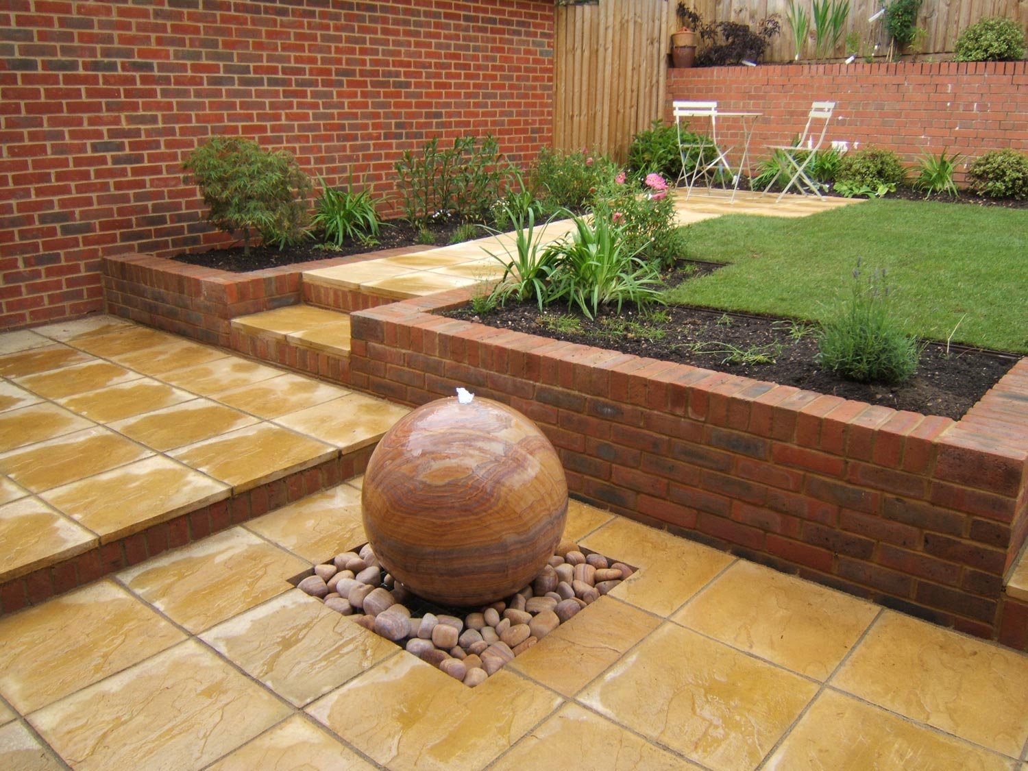 Backyard garden with brick planter beds, a small lawn, a tiled patio, and a water fountain with a round stone sphere.