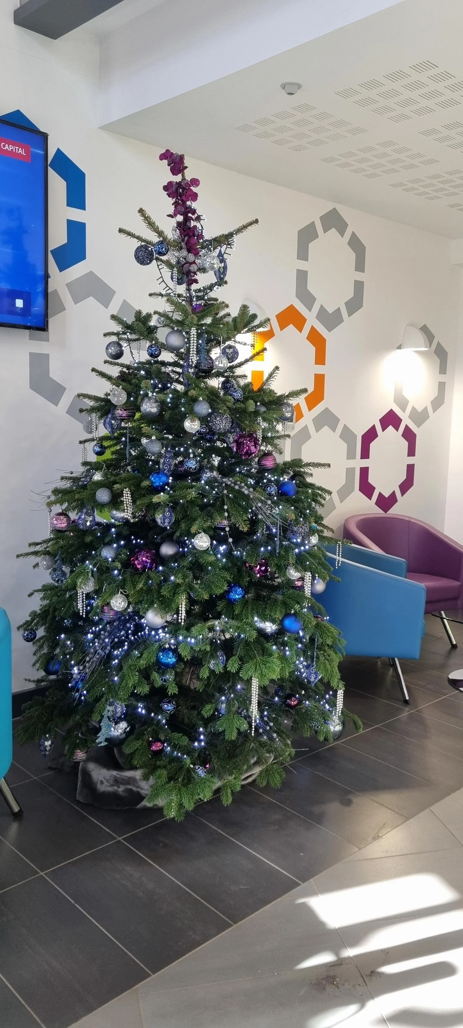 Decorated Christmas tree with silver, blue, and purple ornaments and lights in a modern indoor space.
