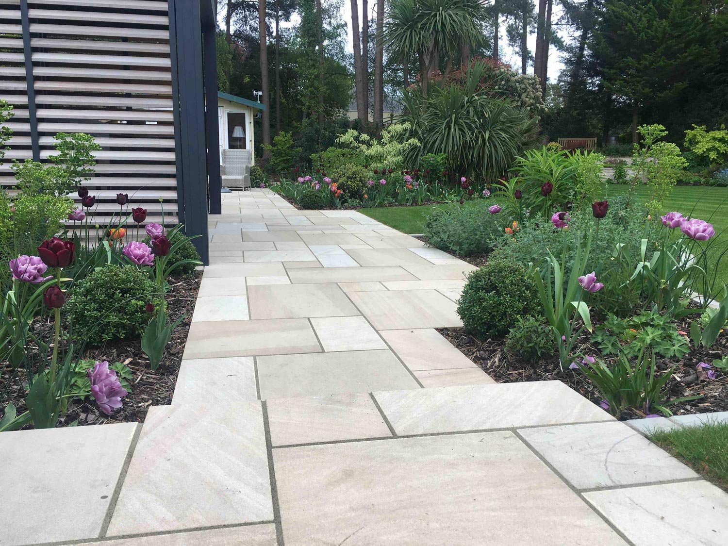 A paved garden pathway with light-colored tiles runs alongside a modern house with horizontal panel siding, surrounded by blooming pink, purple, and dark red tulips and lush green plants, leading to a backyard garden with trees and a small shed in th