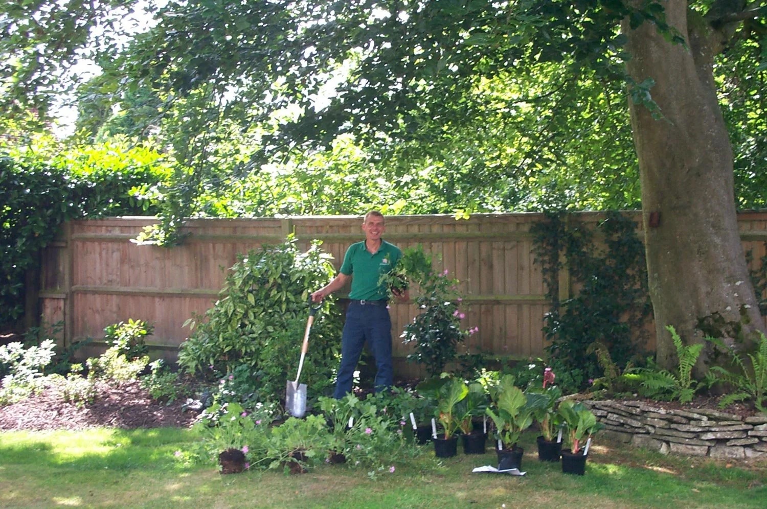 Man gardening in backyard with potted plants, soil, and garden tools, surrounded by trees and a wooden fence.