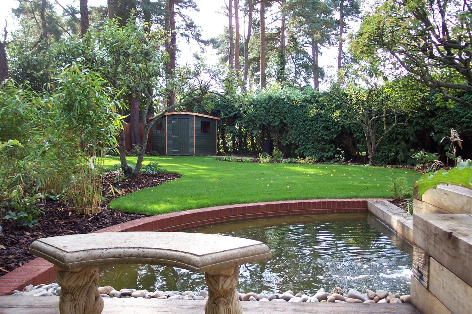 A backyard with a small pond in the foreground, a grassy area, trees, shrubbery, and a wooden shed at the back.