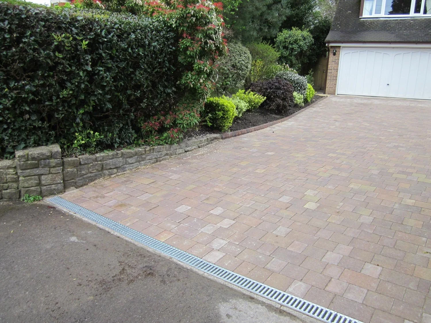 Picture of a paved driveway with a stone border, garden bushes and a garage door in the background.