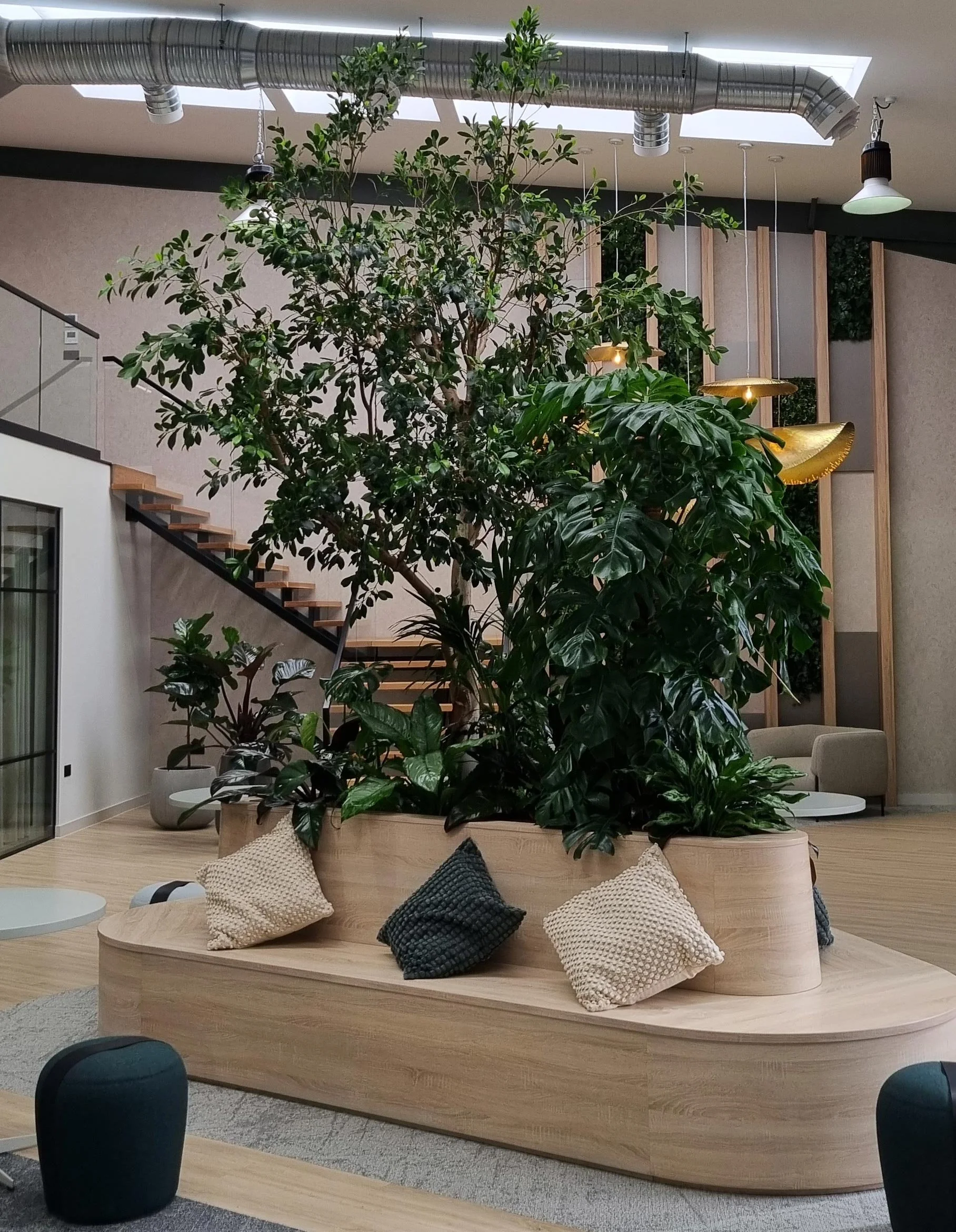 Indoor space with a large central tree in a wooden planter, surrounded by pillows, modern seating, and decorative lighting fixtures, with a staircase and industrial-style ceiling ducting visible.
