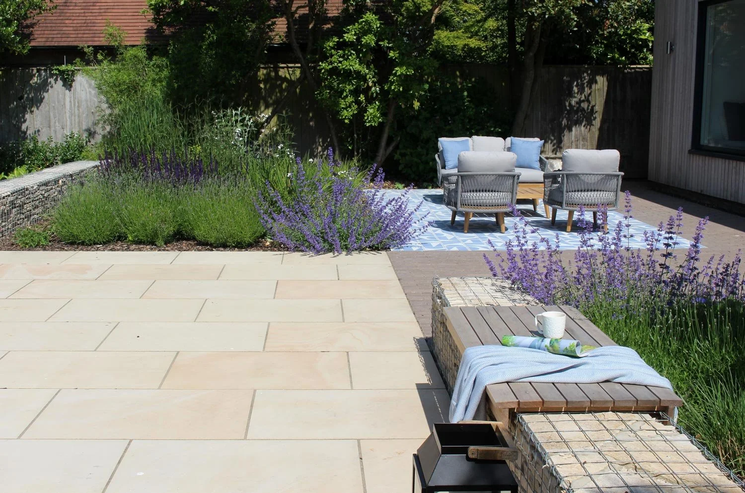 A backyard patio area with outdoor furniture, lavender plants, and a tiled rug, surrounded by greenery and a wooden fence.