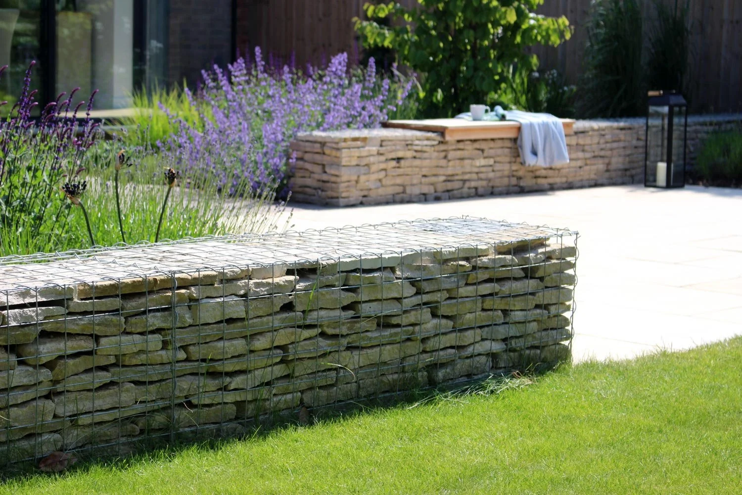 A landscaped backyard with a stone and wire gabion wall, purple flowering plants, a stone bench with a white towel and cups, a black lantern with a white candle, and a deck with potted plants and trees.