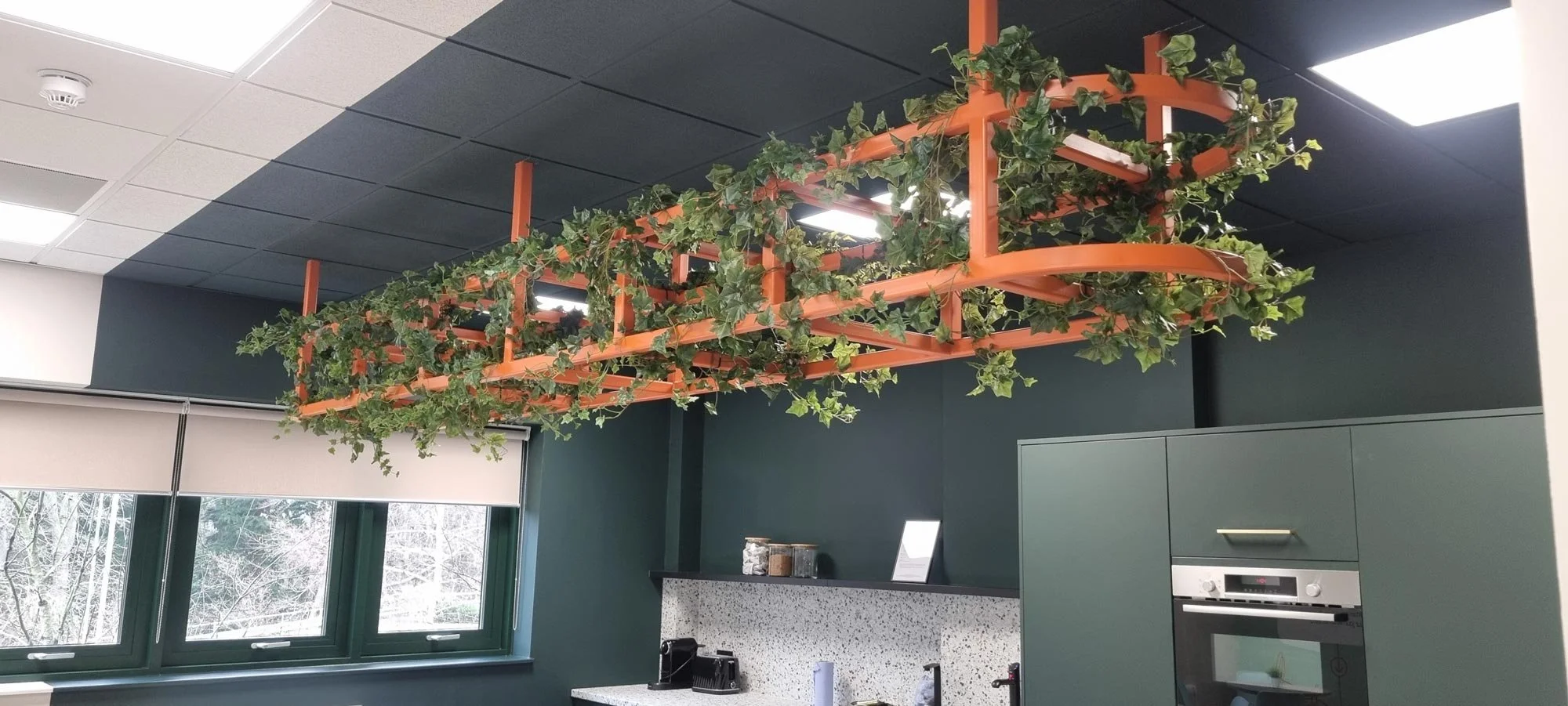 Indoor kitchen area with a ceiling-mounted orange plant holder with green ivy plants, a window showing snow-covered trees outside, and kitchen appliances on a speckled countertop.