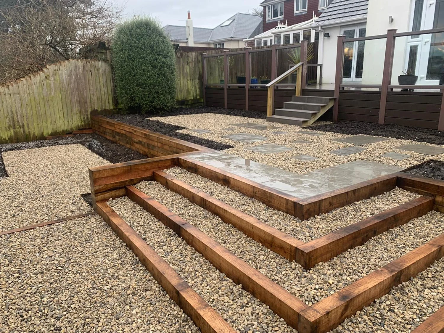 Backyard garden with wooden raised beds, gravel pathways, and a small set of stairs leading to a deck, fenced with wooden panels.