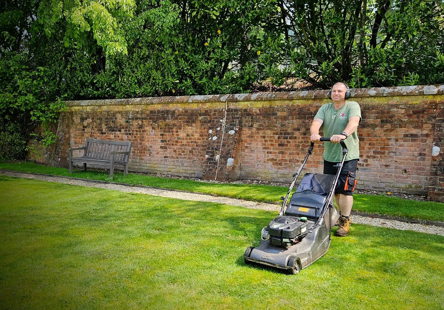 Man mowing lawn with a push lawn mower, wearing headphones, shorts, green T-shirt, and boots, in a backyard with a brick wall and green trees in the background.
