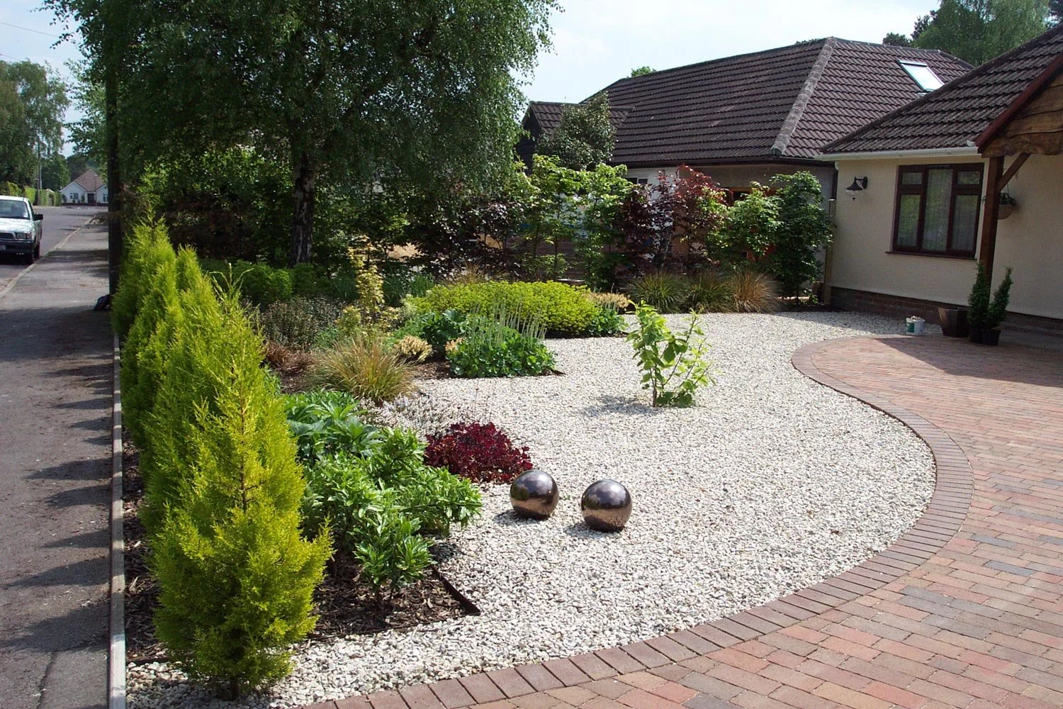 A residential front yard with a curved brick walkway, white gravel ground cover, various green shrubs and plants, small trees, two shiny metallic spheres, and a house with a tiled roof in the background.