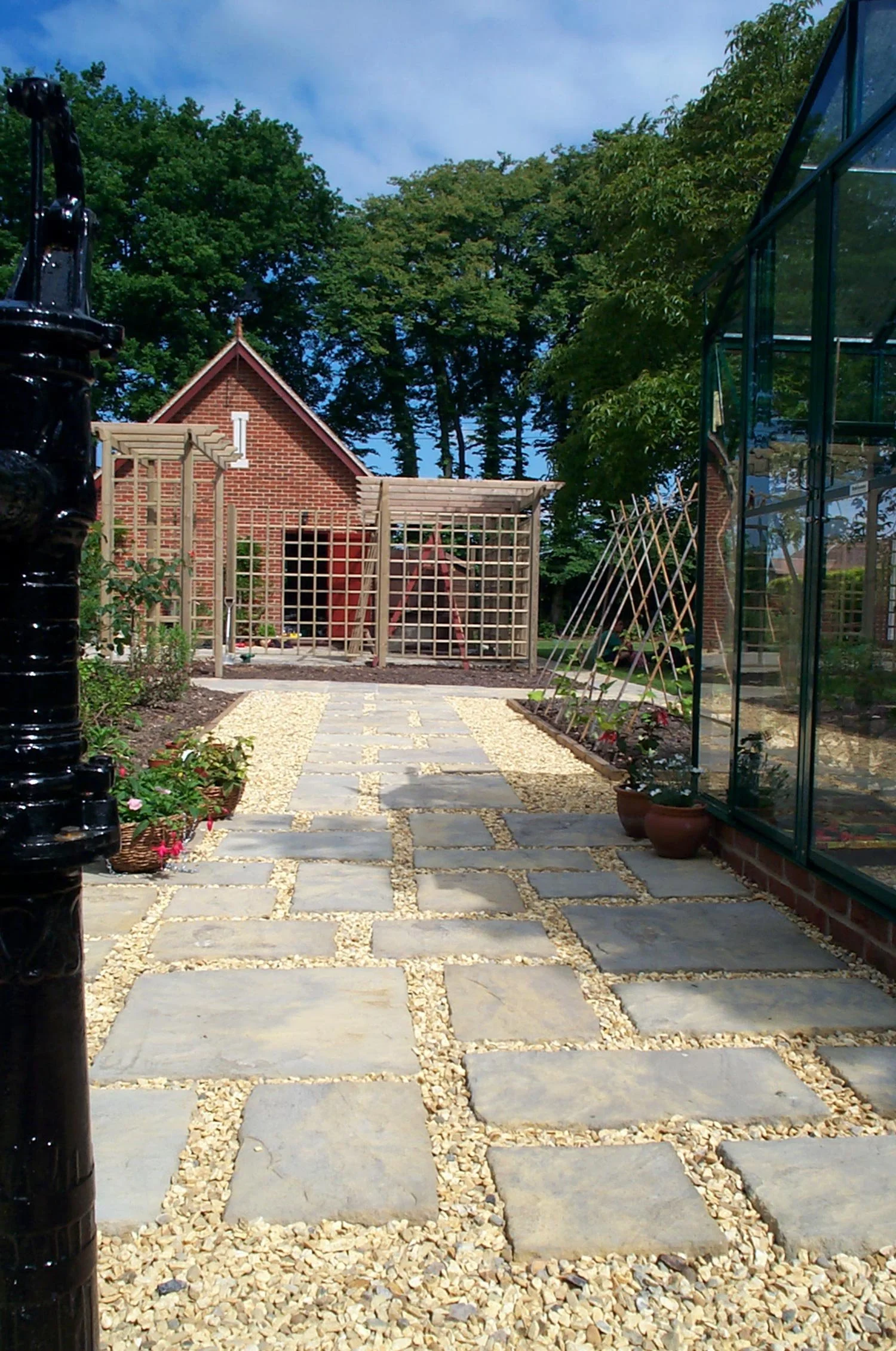 A garden pathway made of large stone slabs and small gravel leads to a small wooden trellis with a brick house and trees in the background. There are potted plants and a greenhouse on the right.