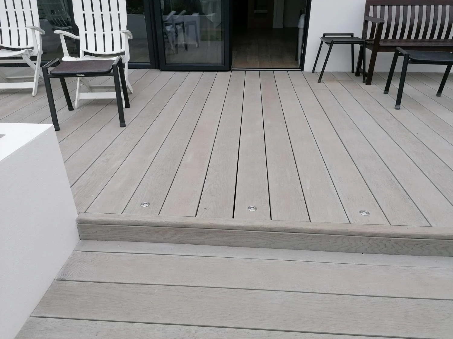 A patio with light gray wooden decking, white and dark brown outdoor chairs, and a glass sliding door leading indoors.