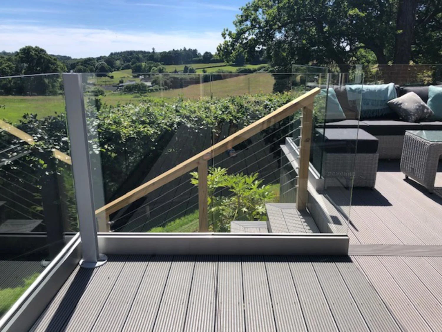 View from a balcony with deck flooring, glass railing, outdoor seating with cushions, and a scenic green landscape with rolling hills and trees under a partly cloudy sky.