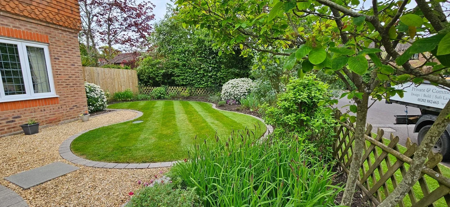 Well-maintained garden with a curved grass lawn, surrounded by various plants and shrubs, a brick house with a window on the left, and a wooden fence with trees on the right.