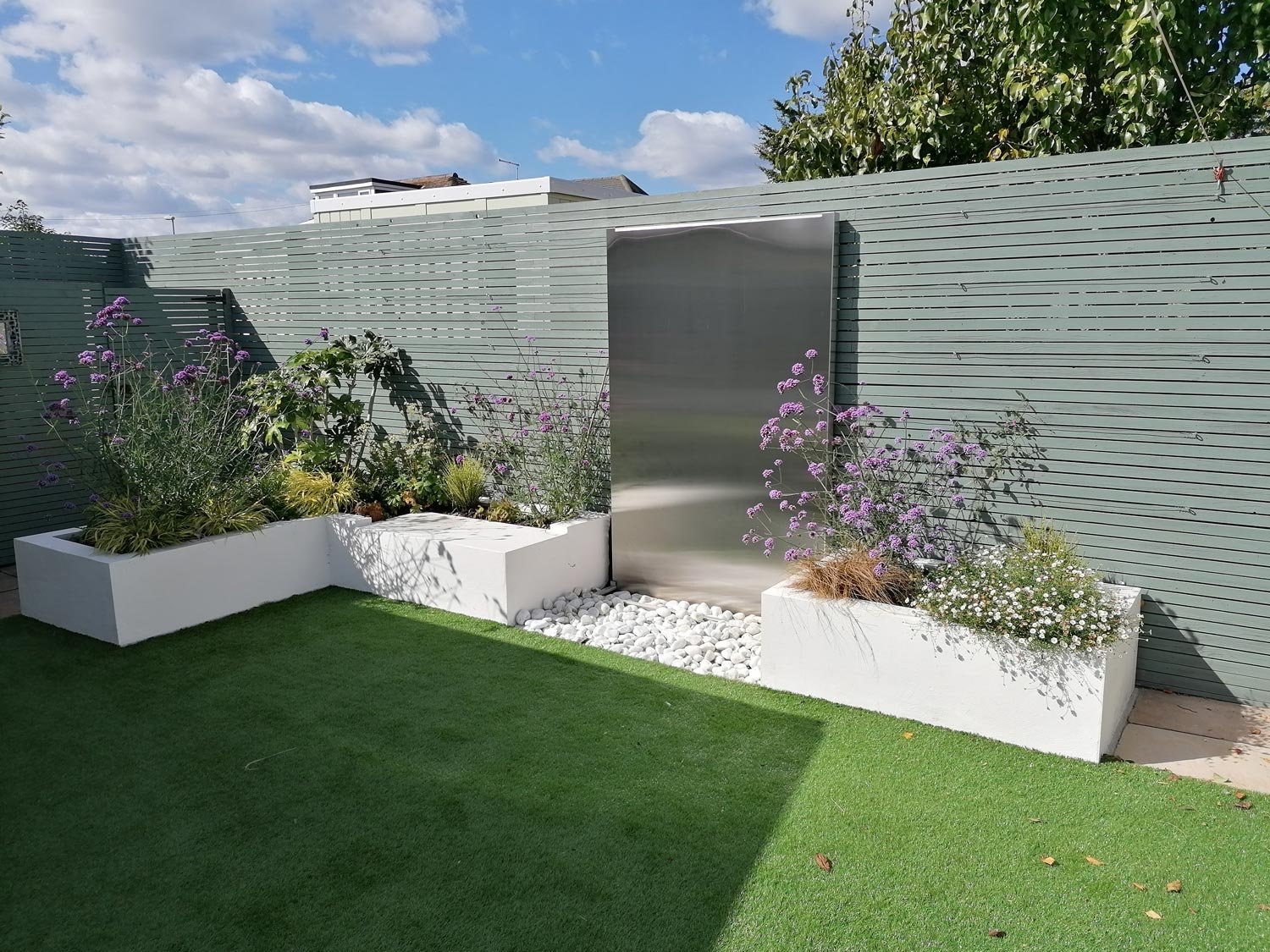 A modern garden with white rectangular planters filled with purple and white flowers, a green lawn, a slatted privacy fence, and a stainless steel water feature under a partly cloudy sky.