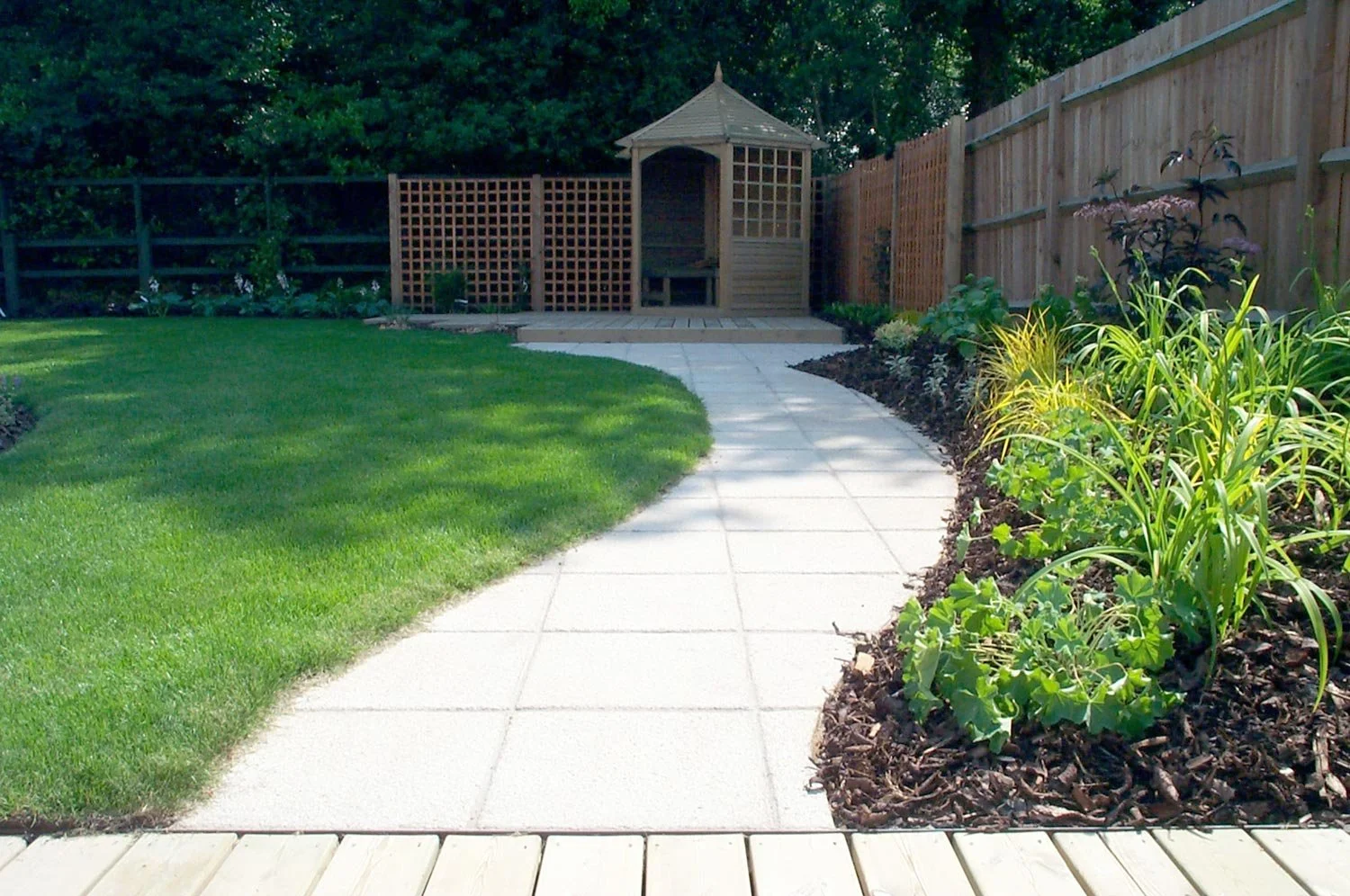 A backyard with a concrete pathway, green grass, and a garden bed with plants on the right. In the background, there is a small wooden shed with a hexagonal roof and a lattice fence.