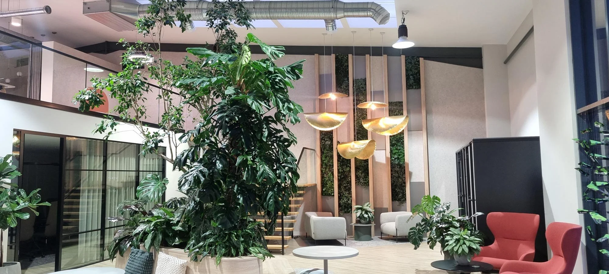 Multiple floors of a building viewed from the ground floor, looking up through an open atrium with a staircase and green plants hanging along the edges of the balconies.