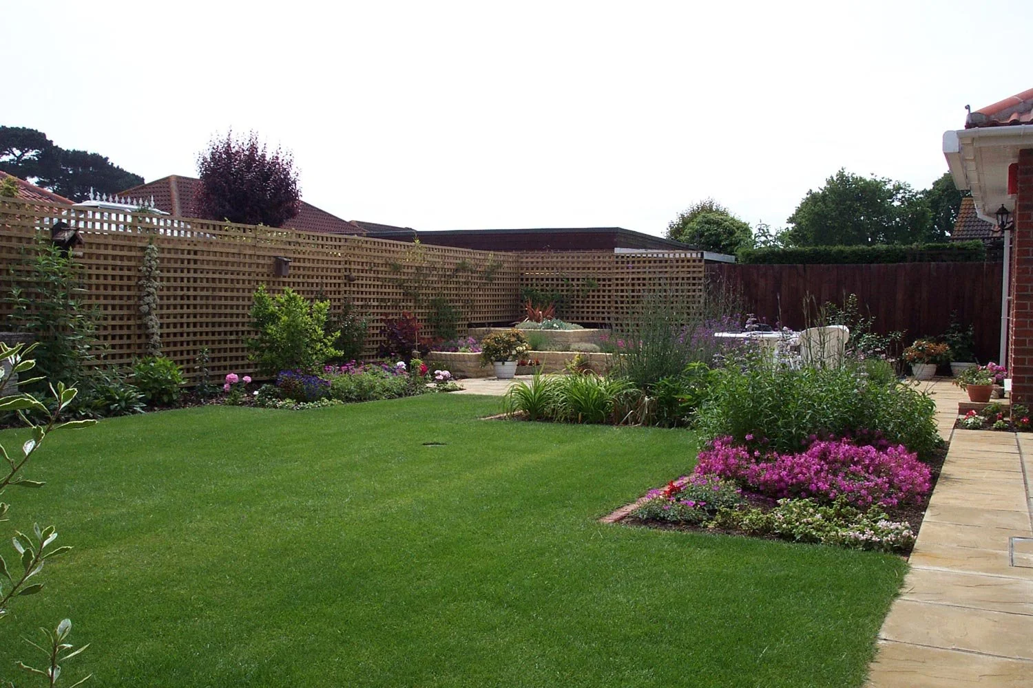 A well-maintained backyard garden with a lush green lawn, flower beds with pink and purple flowers, a curved stone pathway, and a wooden lattice fence.