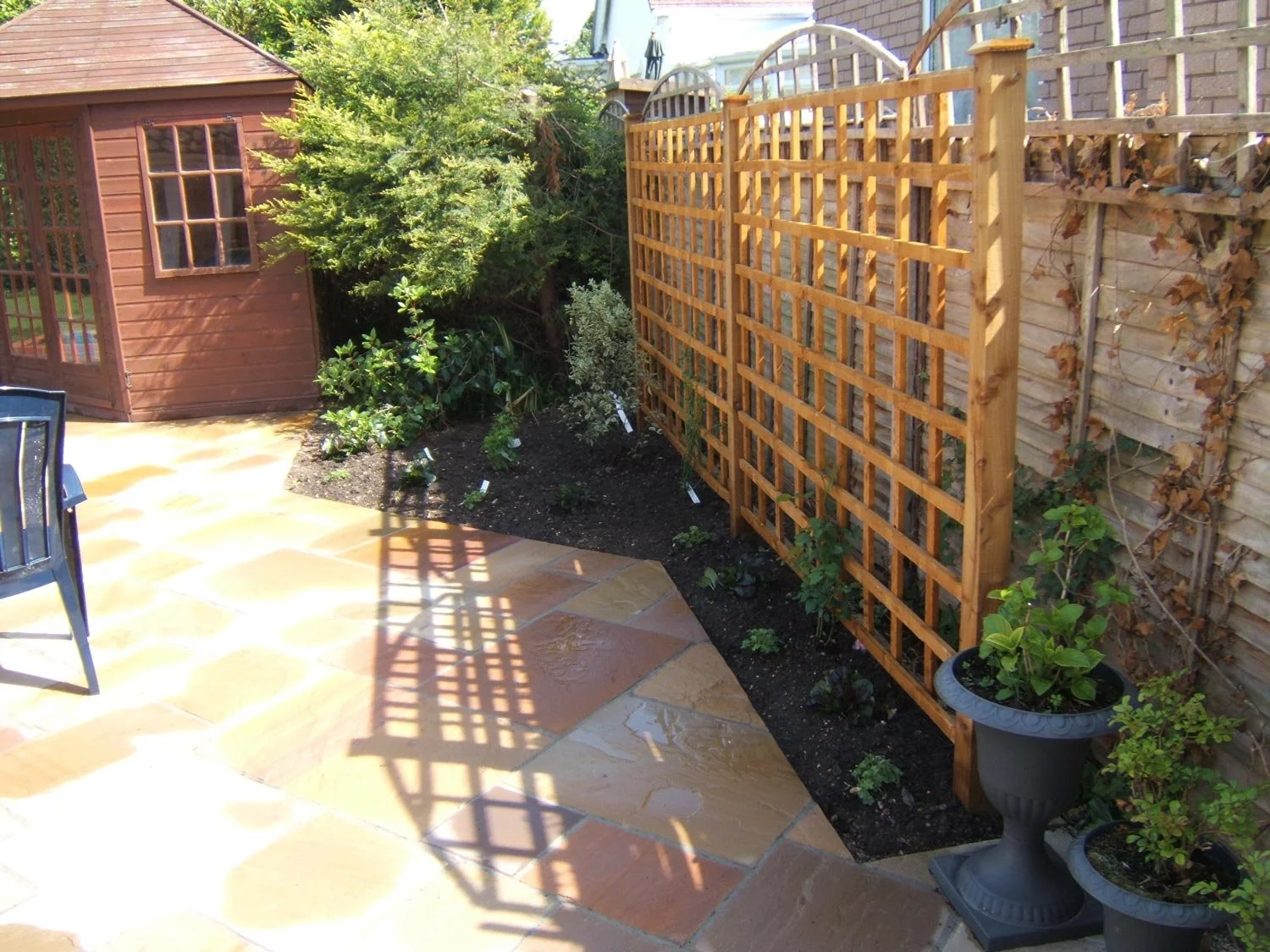 A backyard garden with a paved stone patio, a wooden shed, a trellis with plants, and potted plants along a wooden fence.