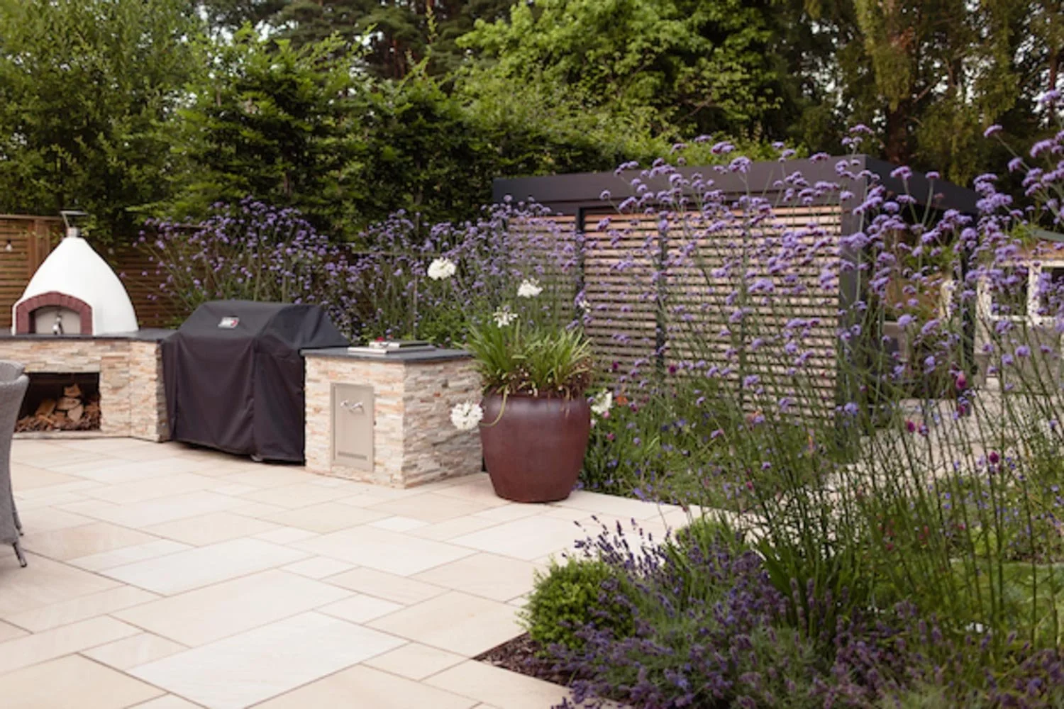 A backyard patio featuring a grill under a cover, a wood-fired oven, and large potted plants with purple flowers. The area is enclosed by a wooden fence and surrounded by greenery and flowering bushes.