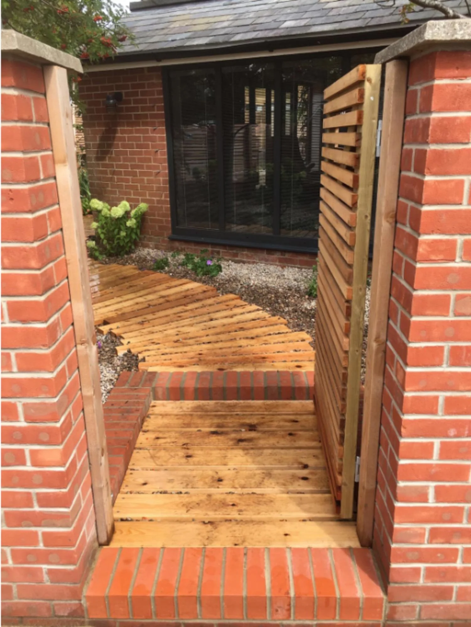View of a brick porch with a new wooden deck installation and a white brick house with a large window.