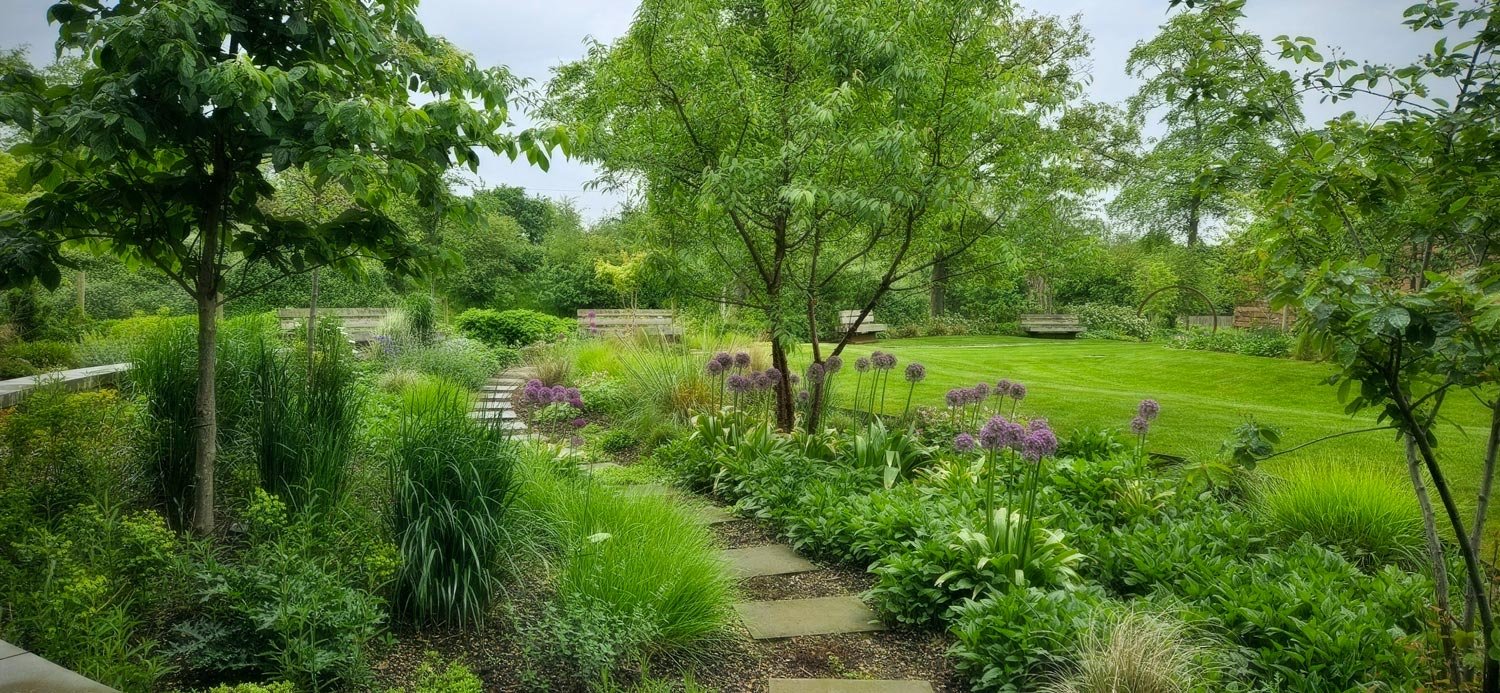 A lush garden with a stone pathway, green grass, trees, and blooming purple flowers.