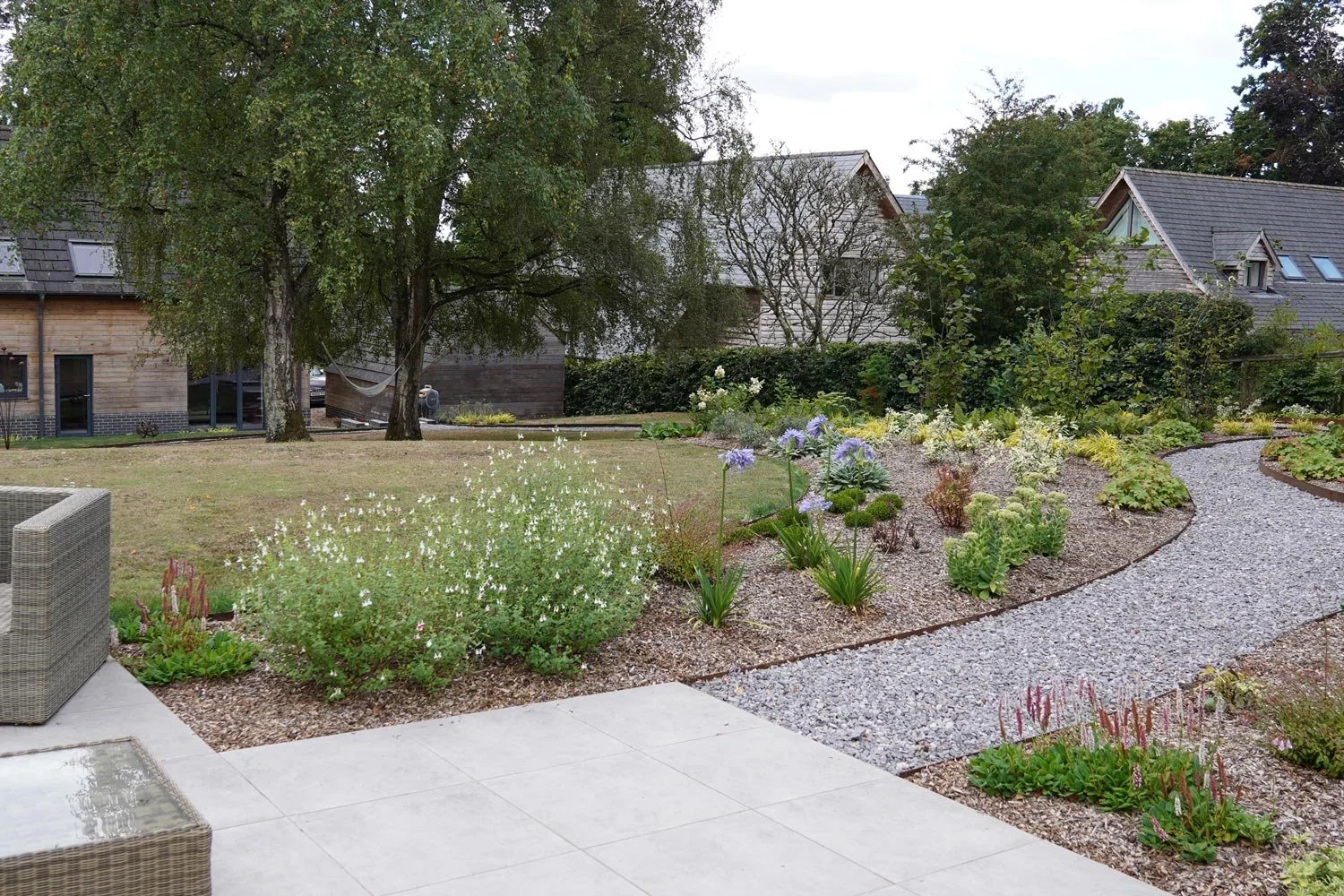 Backyard garden with trees, flowering plants, and a gravel pathway, adjacent to a patio with outdoor furniture, modern houses in the background.