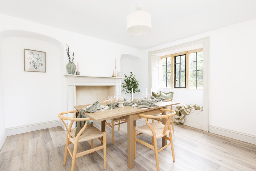 Bright dining room with a wooden table, six chairs, a window seat, and a fireplace with decorative items, all in a minimalist style.