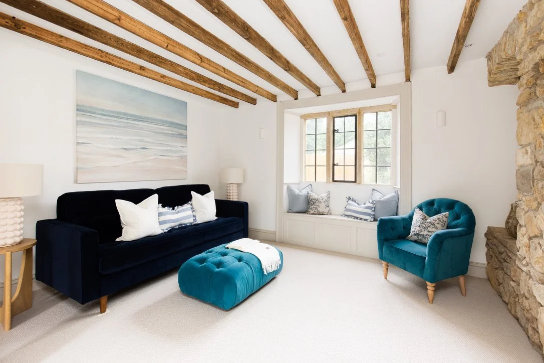Living room with white walls, wooden ceiling beams, a dark blue sofa, a matching blue armchair, a window seat with pillows, a stone wall section, and a large landscape painting.