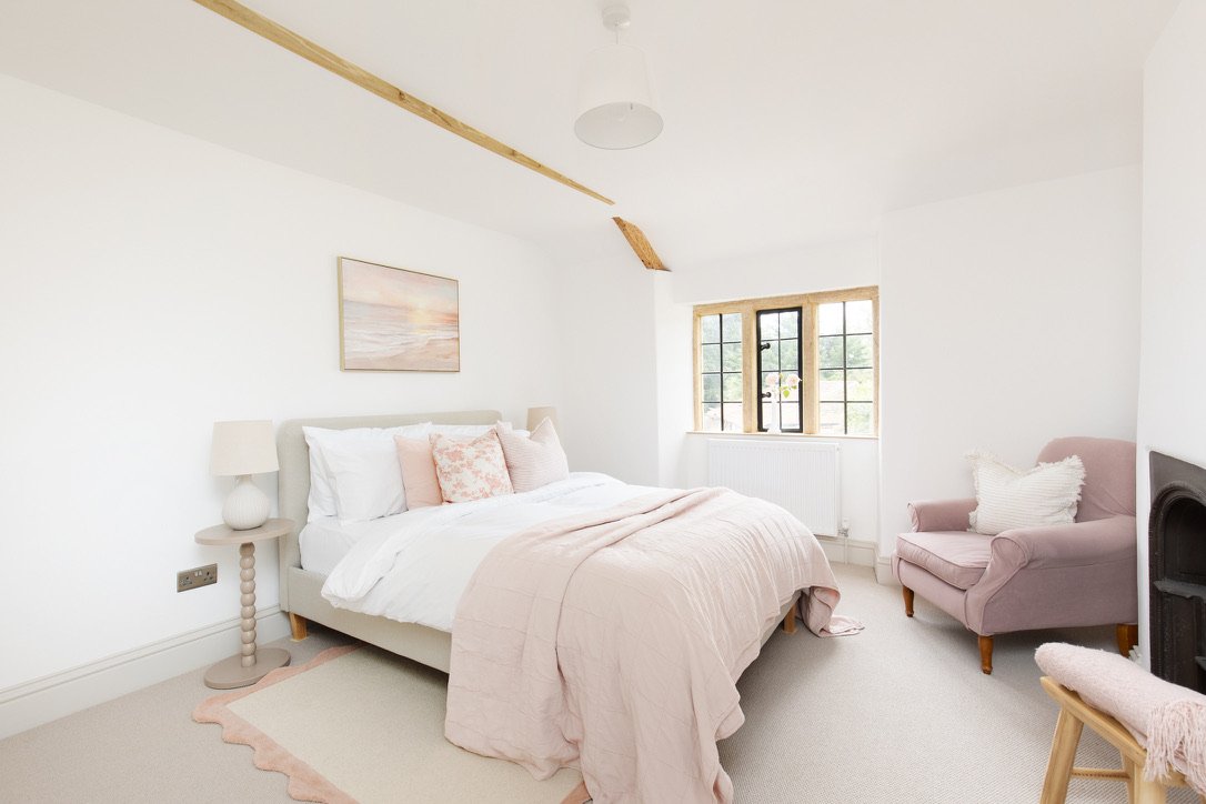A bright bedroom with a white bed, pink bedding, a pink armchair, and a window with a view of greenery.