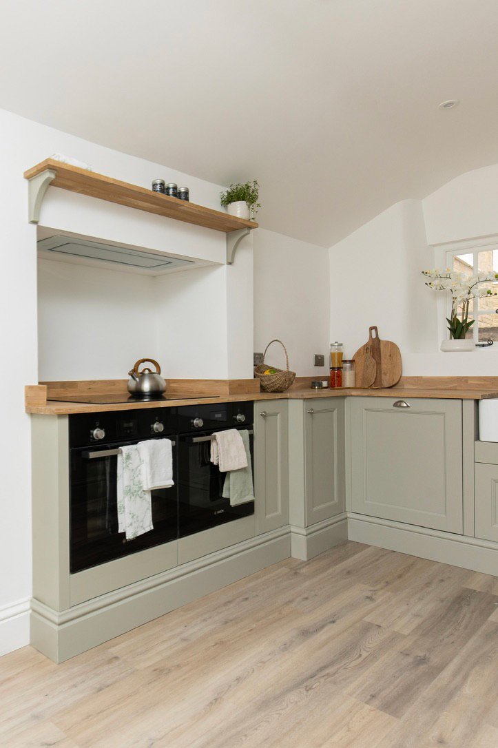 Kitchen with a black oven, wooden countertops, light green cabinets, a small window with a orchid plant, and decorative jars and cutting board on the countertop.
